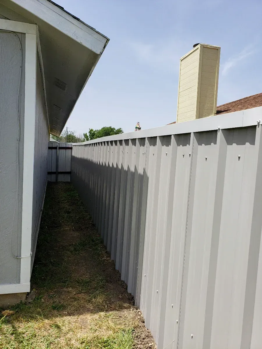 Gray metal fence runs along a narrow grassy space beside a gray house wall and chimney.
