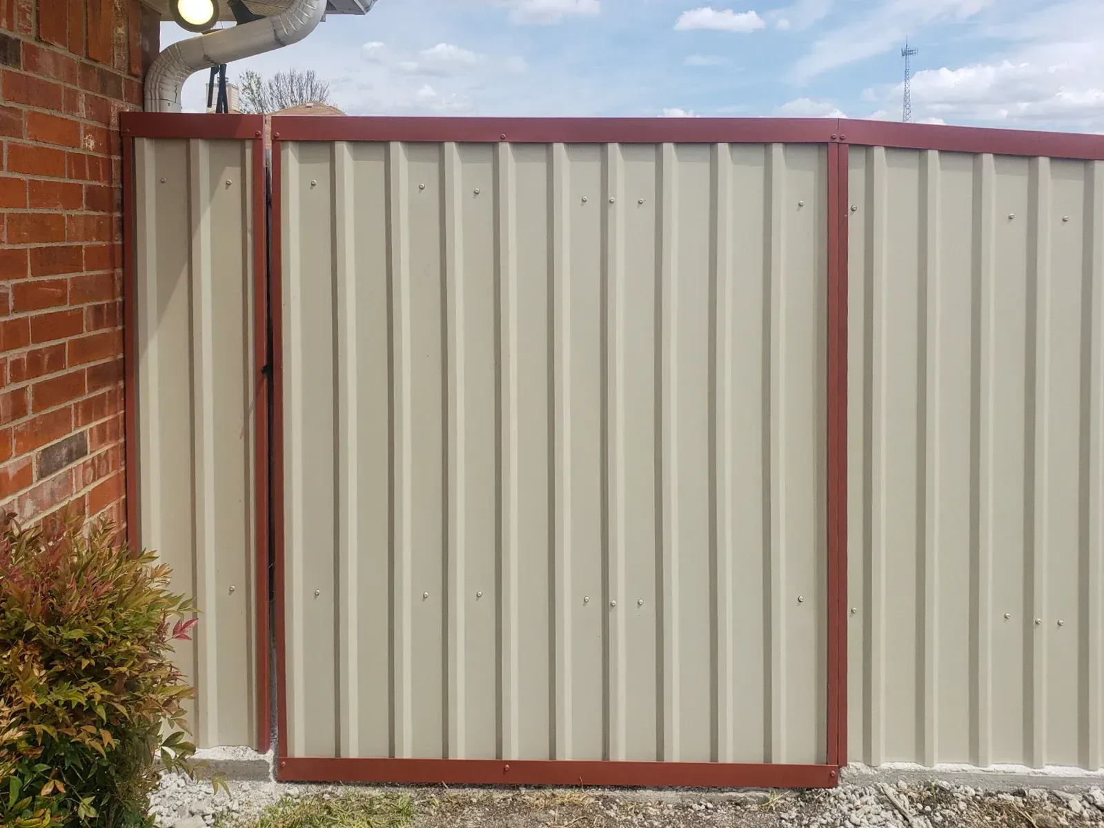 Beige corrugated metal fence with a maroon frame next to a brick wall.