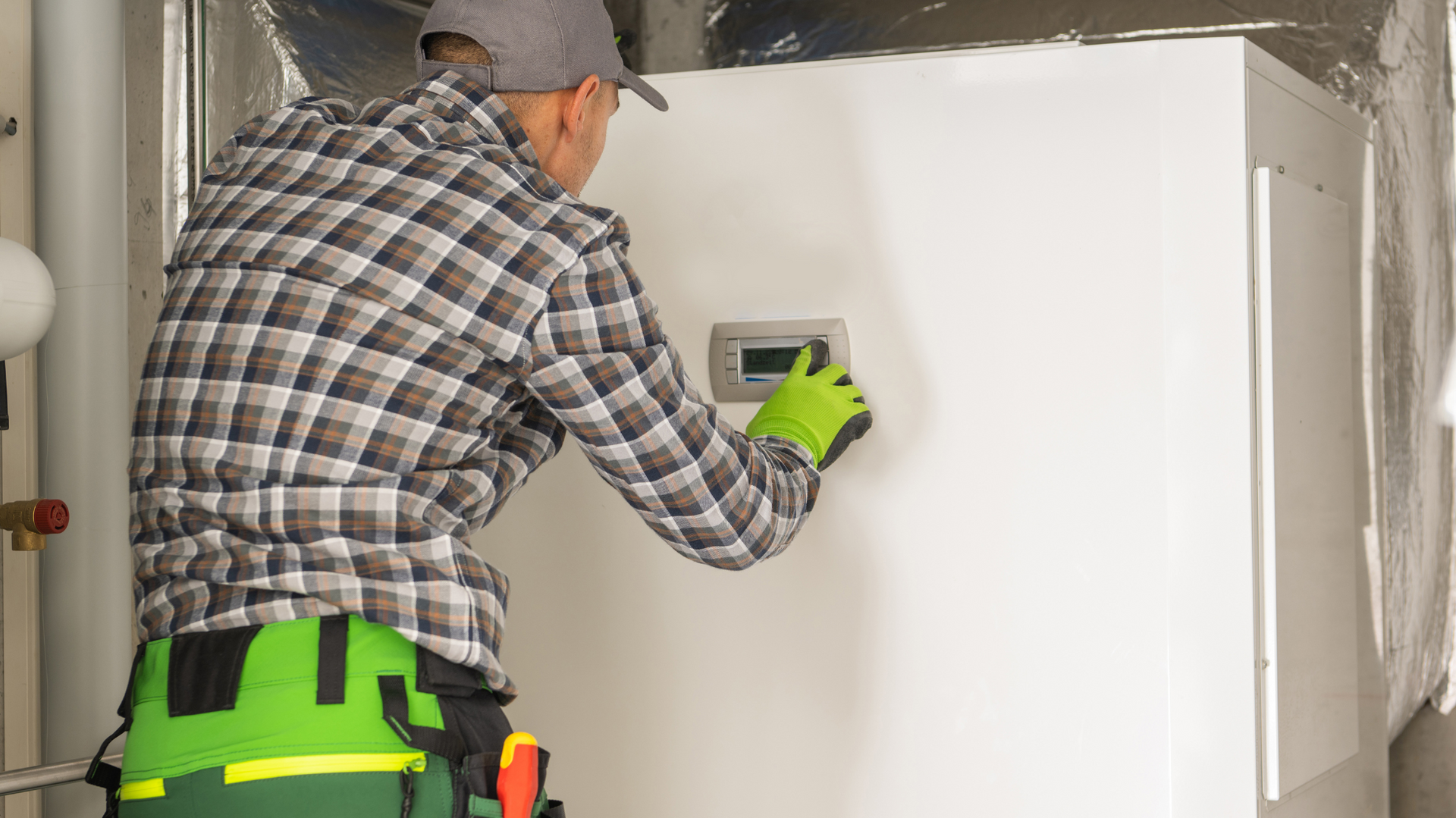 A man in a plaid shirt and green pants is installing a thermostat on a wall.