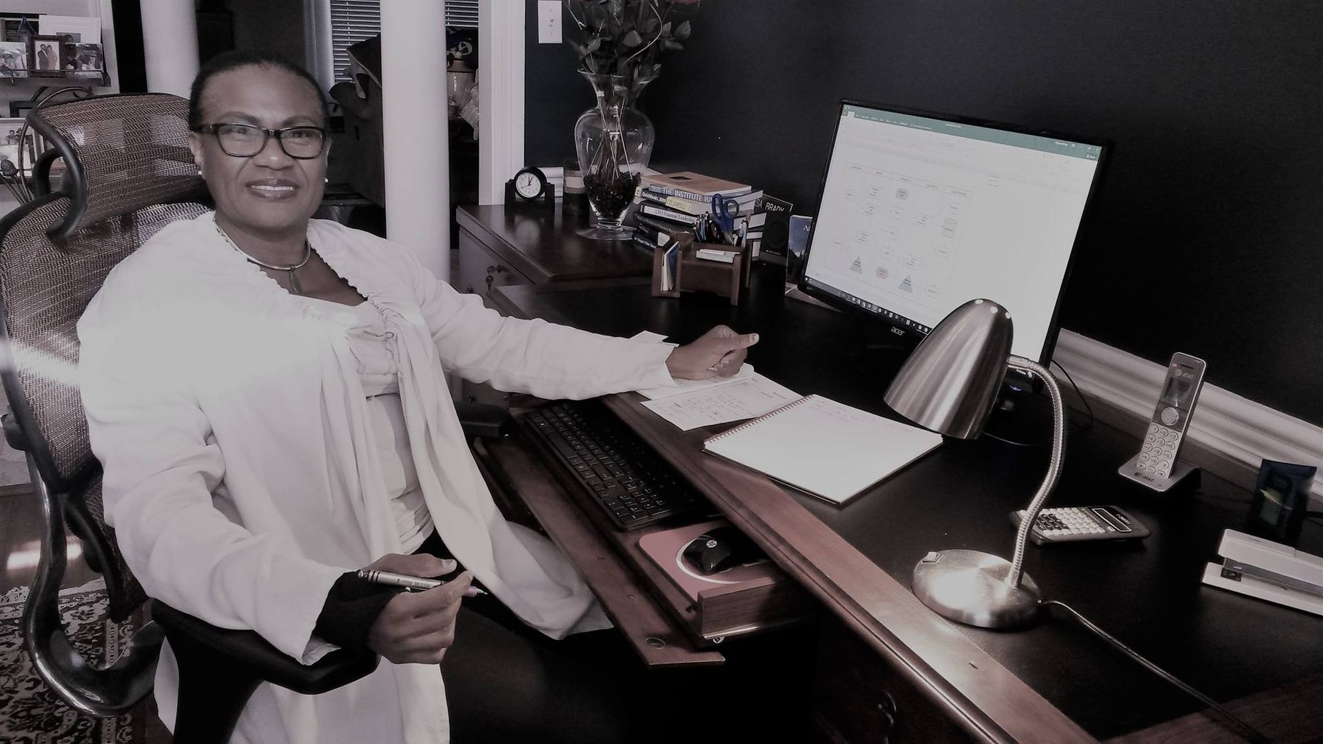 Woman at desk, writing. Computer, lamp, and papers on a dark wooden desk.