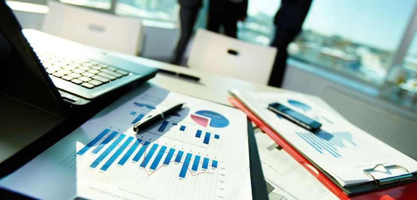 A laptop, charts, and a pen on a table during a meeting with people blurred in the background.