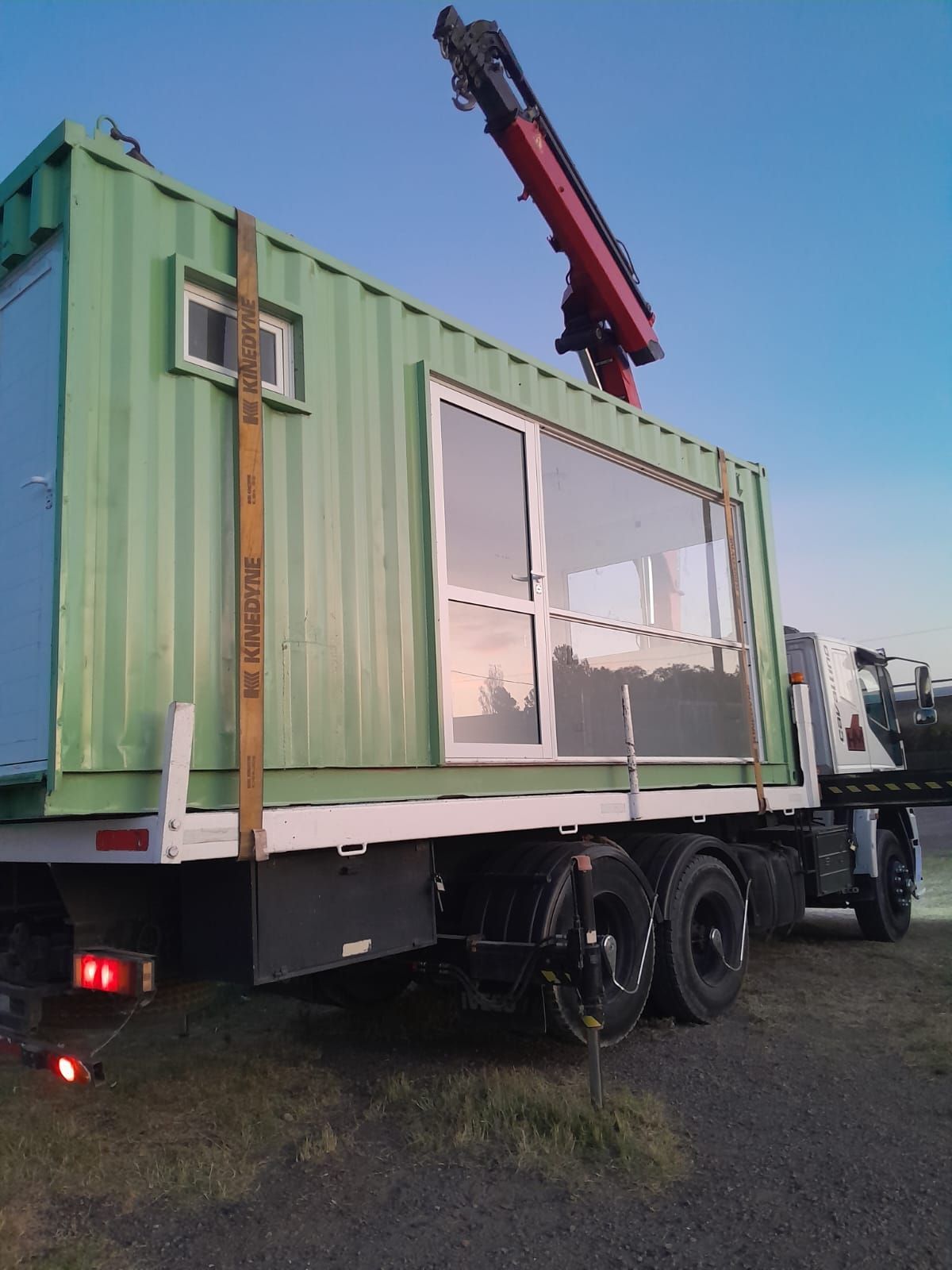 Casa contenedor verde sobre la plataforma de un camión, siendo descargada por una grúa.