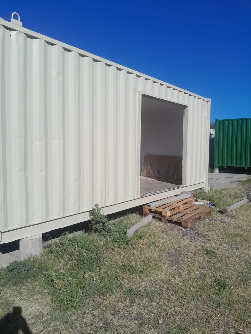 Contenedor de envío de color marrón con una puerta abierta en un terreno cubierto de césped bajo un cielo azul.