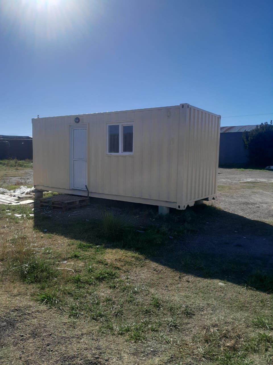 Contenedor de envío de color beige con puerta y ventana, colocado sobre bloques en un entorno al aire libre.