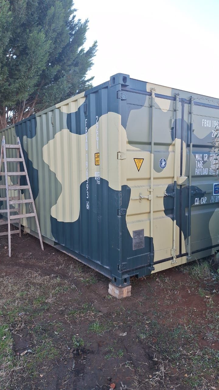 Contenedor de envío pintado con camuflaje, con una escalera de madera a su lado en el exterior.