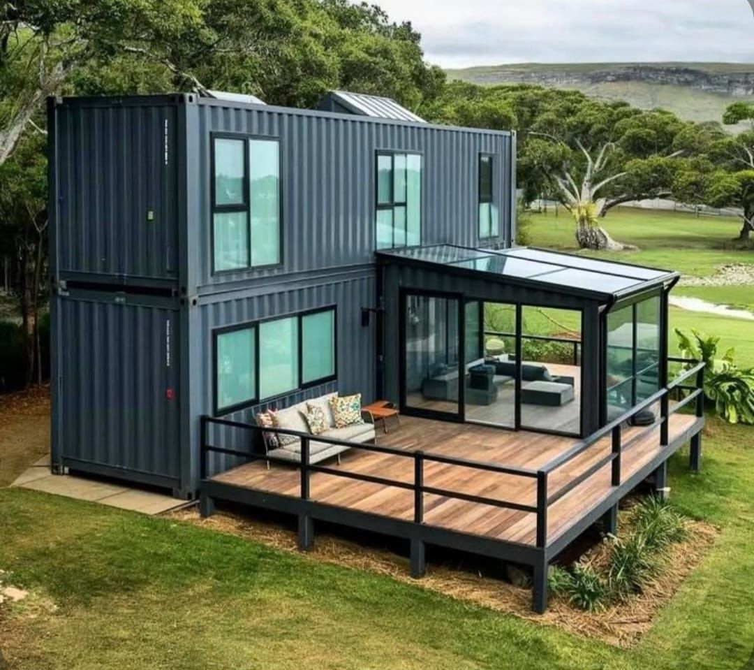 Casa contenedor de dos plantas con terraza, cerramiento de cristal y amplios ventanales. Exterior gris, ubicada en un campo de hierba.