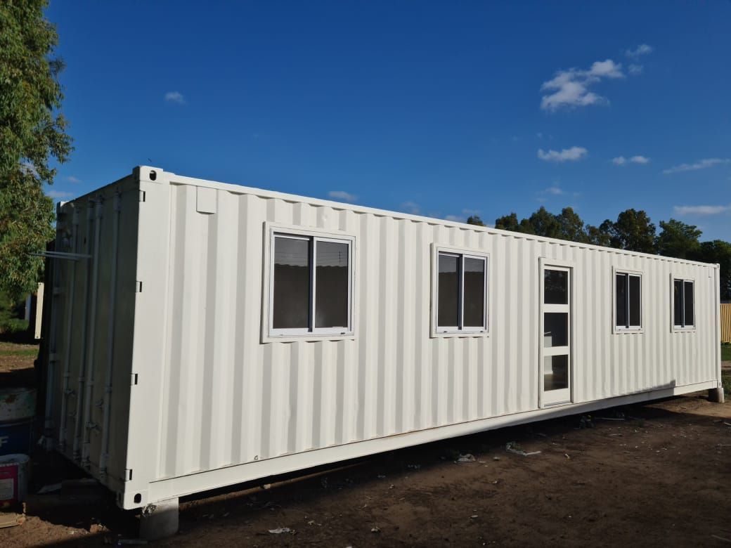 Contenedor de envío blanco modificado con ventanas y una puerta, se encuentra al aire libre en un día soleado.