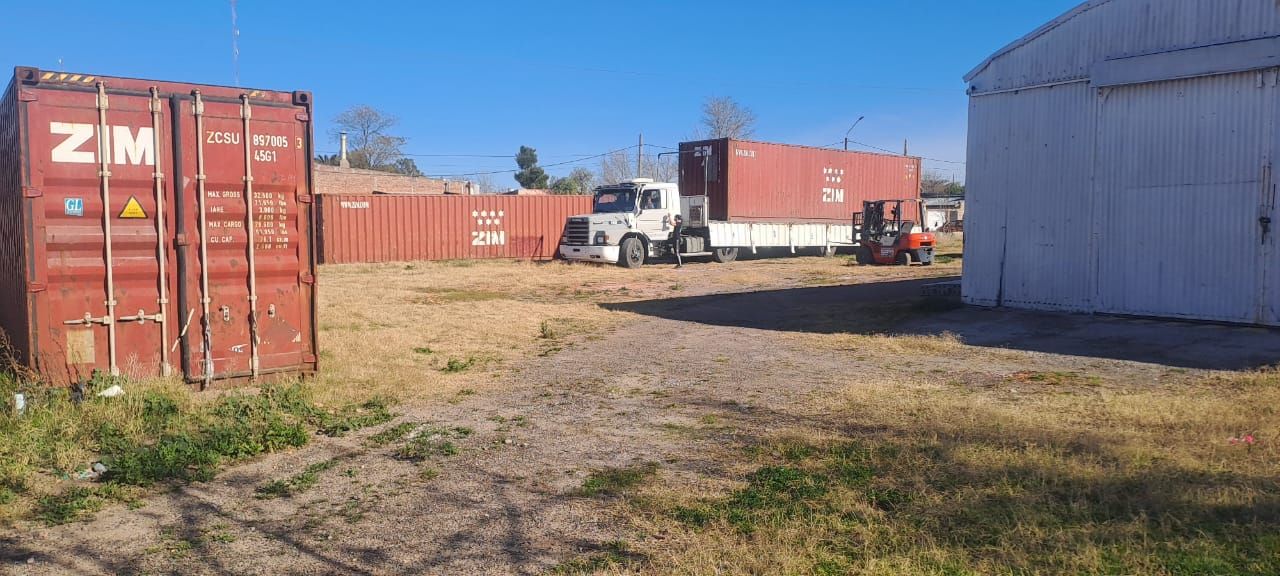 Un camión y contenedores de envío en un lote, con un edificio y un cielo despejado al fondo.