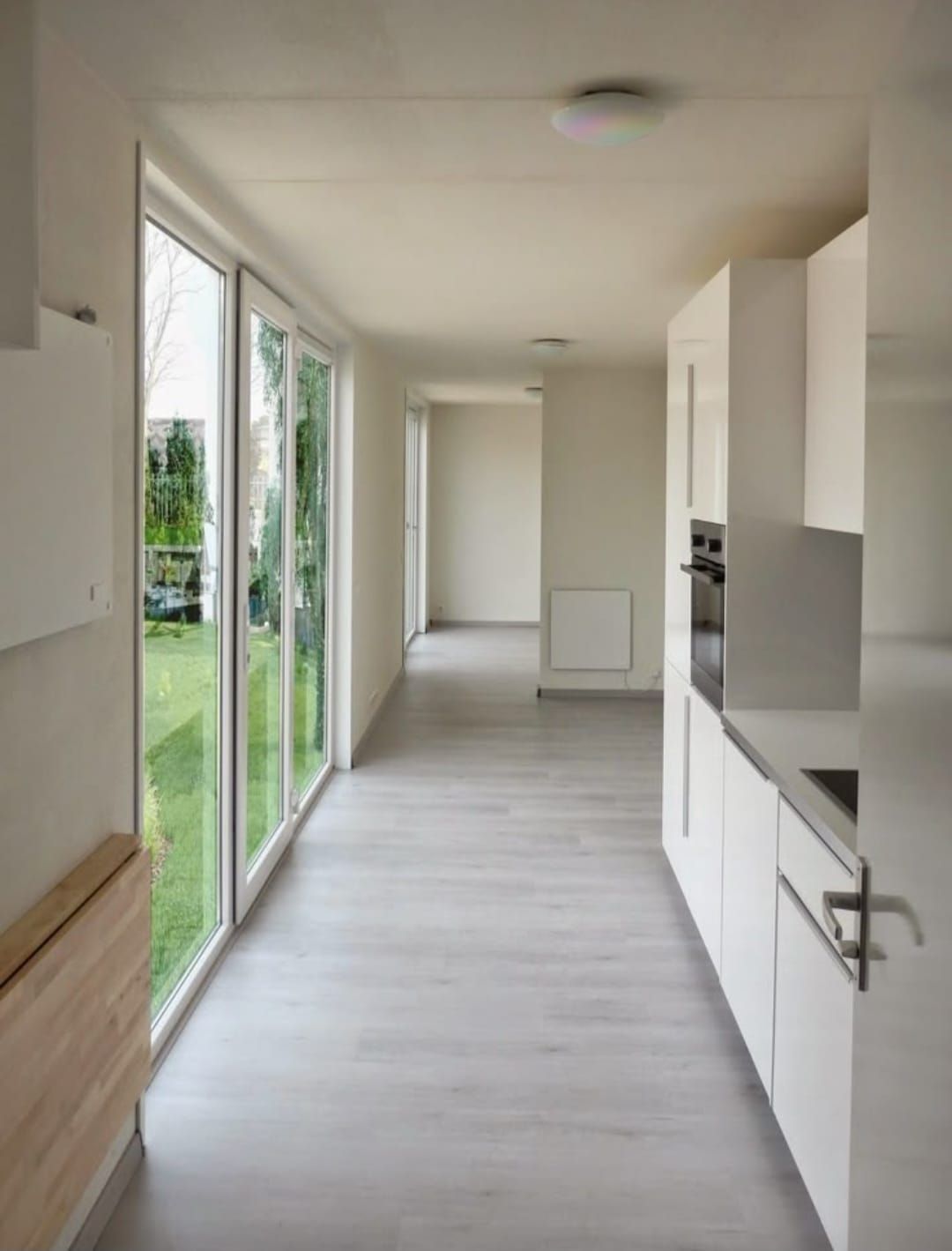 Pasillo largo y luminoso con puertas de vidrio a un patio, gabinetes blancos y pisos de madera clara.