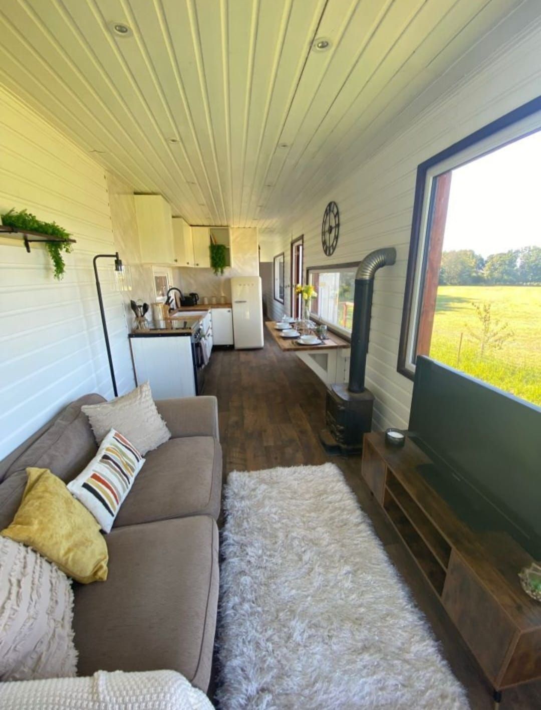 Vista interior de una pequeña y moderna cabaña con sofá, cocina, estufa de leña y gran ventanal con vista a un campo.
