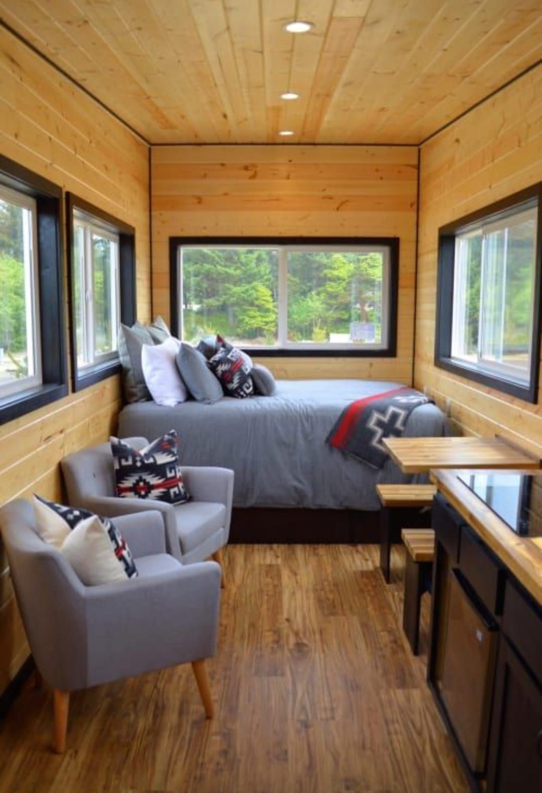 Acogedor interior de casa pequeña: paredes de madera, cama, dos sillones, encimera de cocina y ventanas con vista a la vegetación.