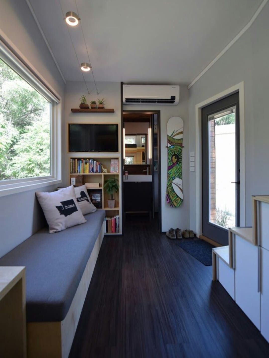 Espacio pequeño y moderno con sofá empotrado, librería, televisor y tabla de snowboard. Suelo oscuro, paredes claras y una ventana.