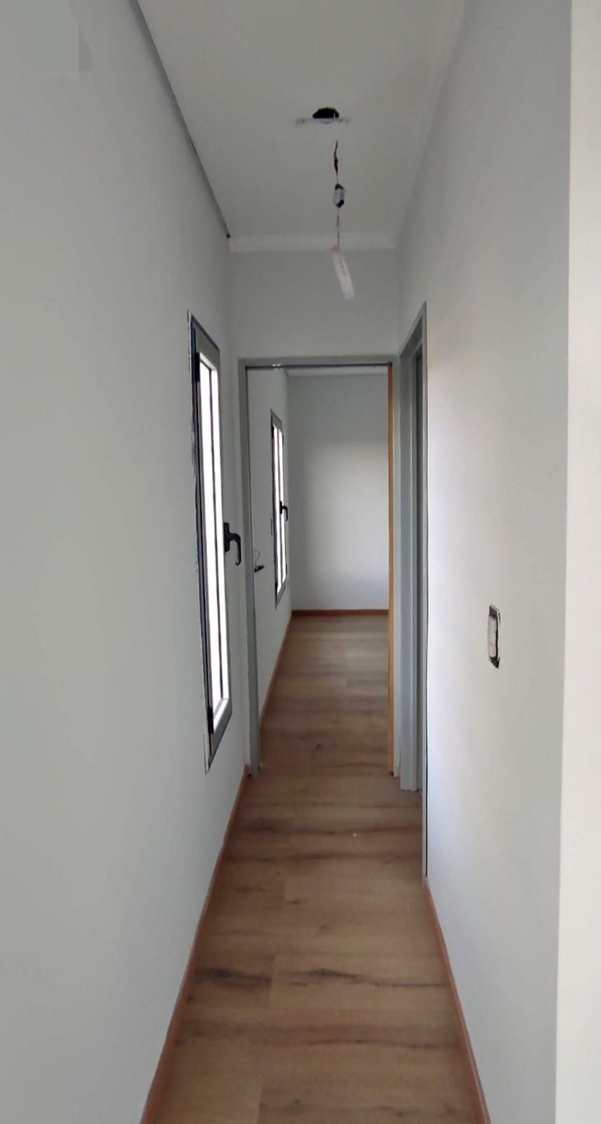 Pasillo estrecho con suelo de madera, paredes blancas, ventanas y puerta abierta.