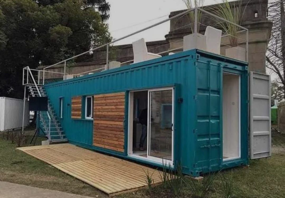 Casa contenedor de envío de color verde azulado con detalles de madera, escalera y patio en la azotea cerca de un edificio.
