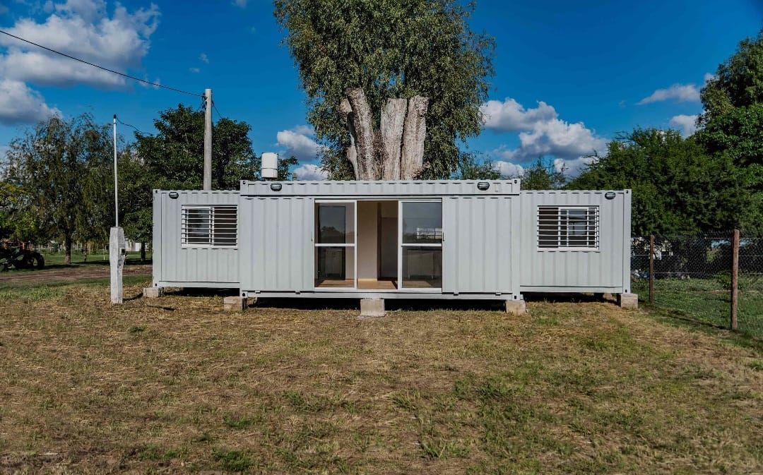 Casa contenedor de envío blanca con ventanas y puertas corredizas sobre un terreno cubierto de hierba bajo un cielo azul.