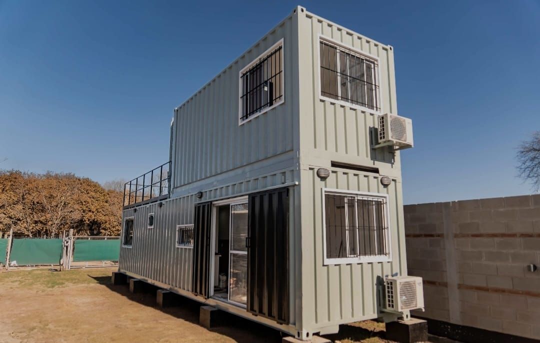 Casa contenedor de dos pisos con puerta abierta, ventanas y unidades de aire acondicionado; ubicada al aire libre bajo un cielo azul claro.