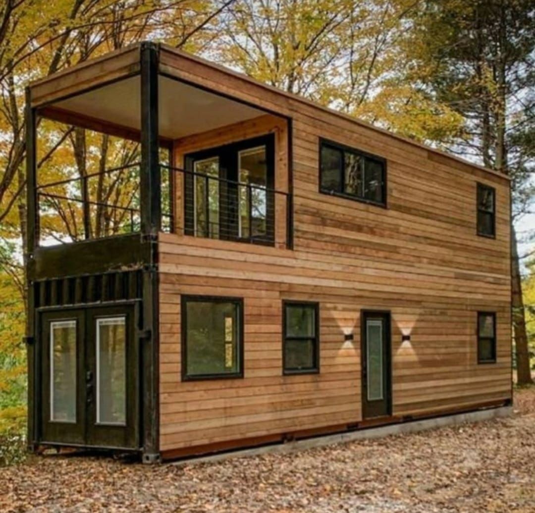 Casa contenedor de envío de dos pisos con revestimiento de madera y balcón en un entorno boscoso.