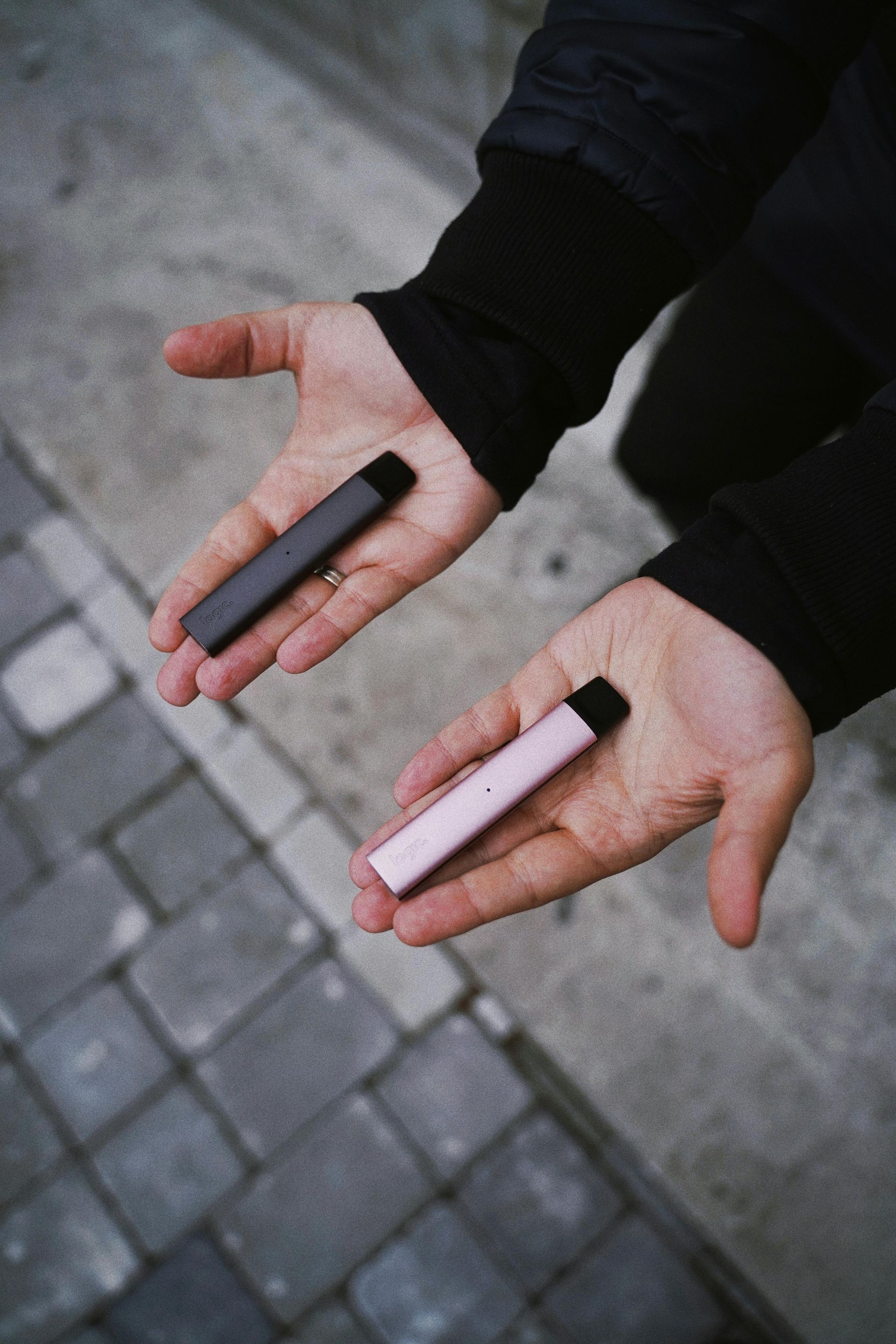 Hands holding a gray and pink vaping device, set against a brick and pavement background.