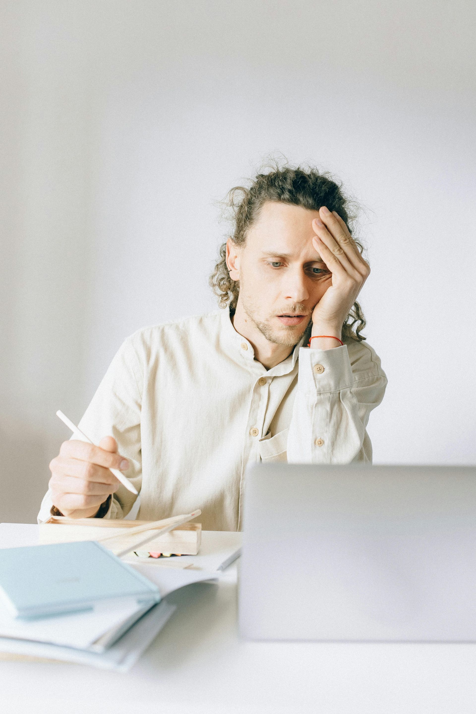 Man with curly hair looks at laptop with a stressed expression, holding a pen. Papers, notebook on desk.