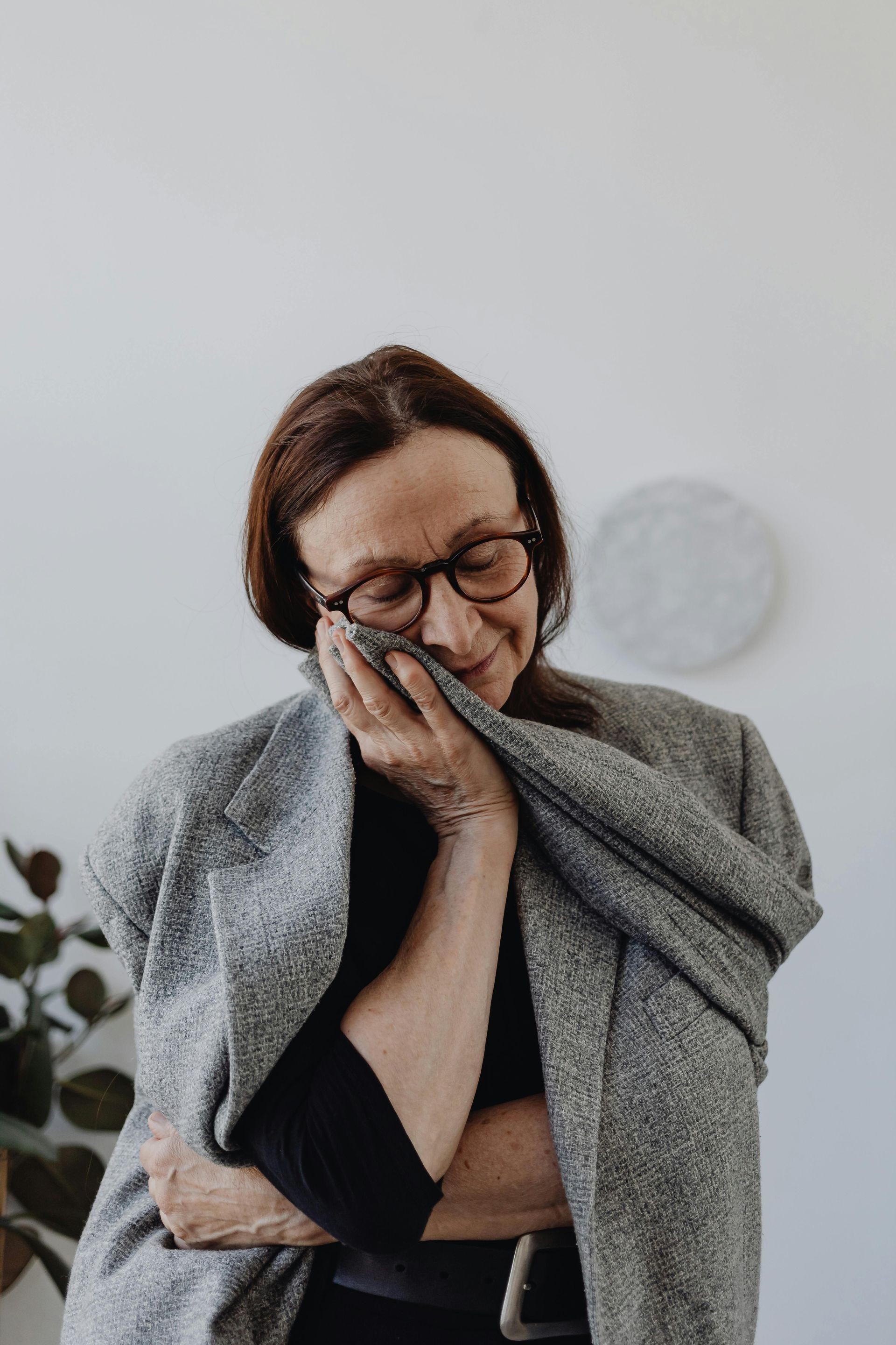 Woman in glasses holding a fabric to her face, smiling. Wearing a gray jacket, standing indoors near a plant.