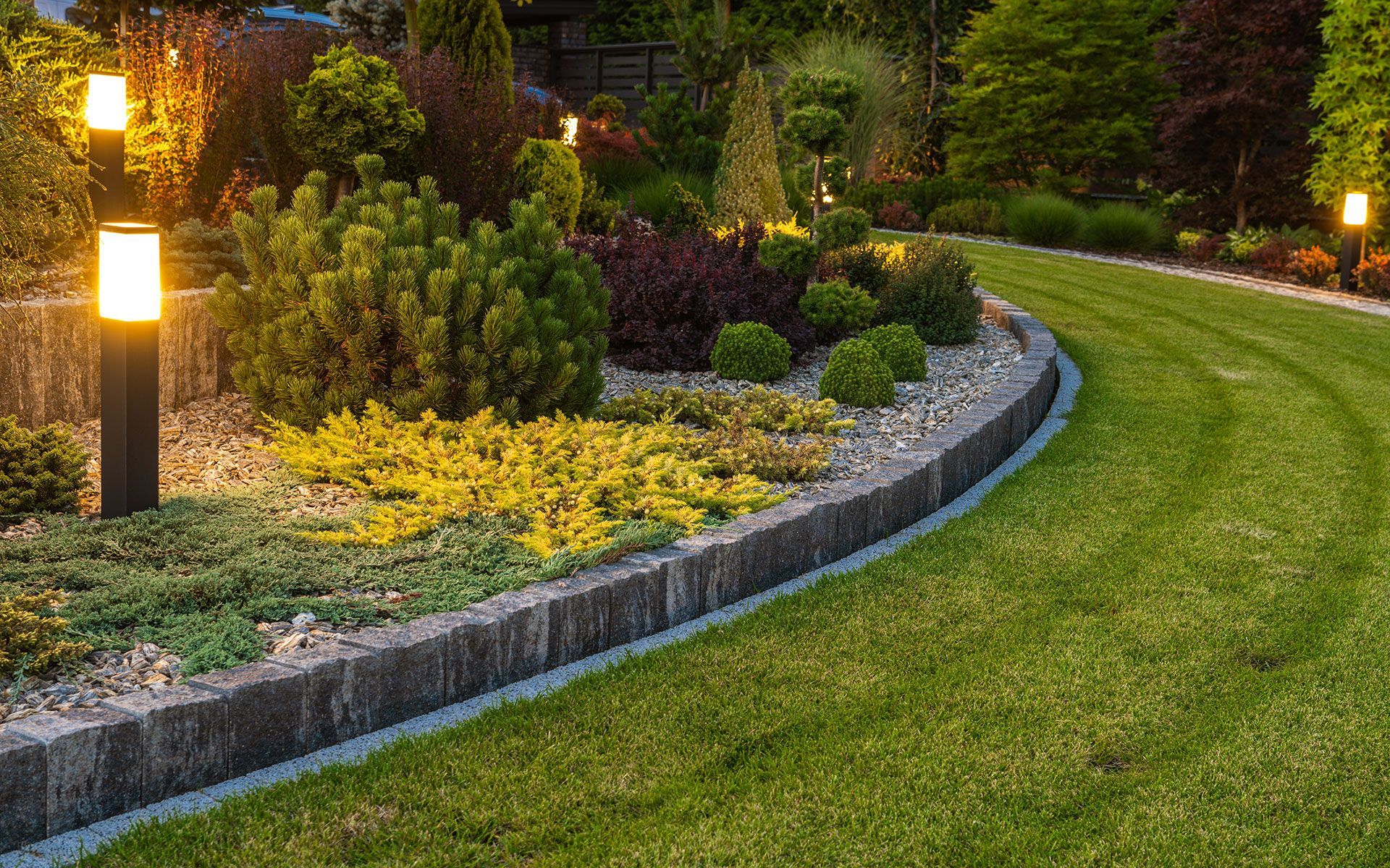 A garden bed with various plants and low lighting, bordered by a stone wall and green lawn.
