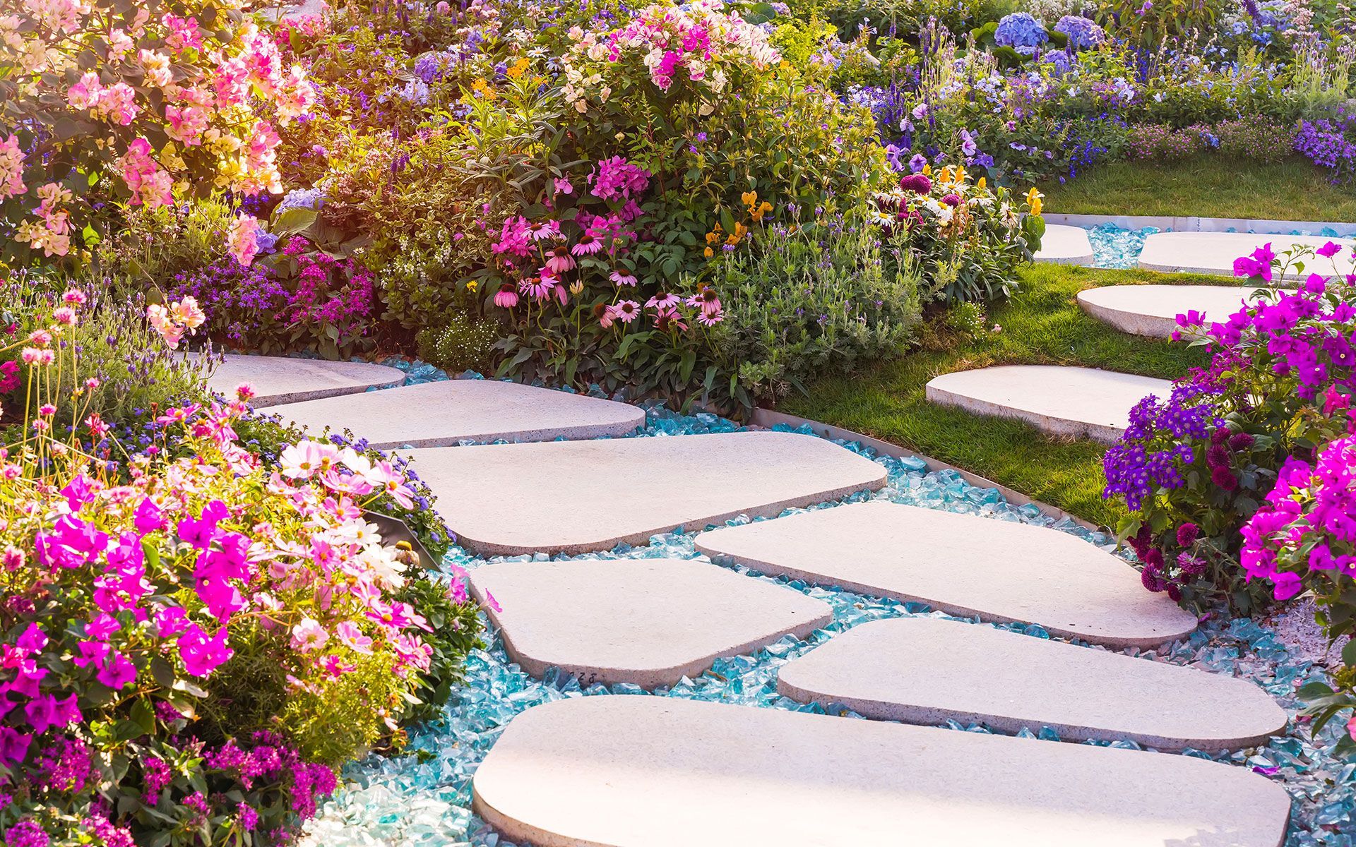 Stone path winds through a vibrant garden with pink and purple flowers, blue pebbles.