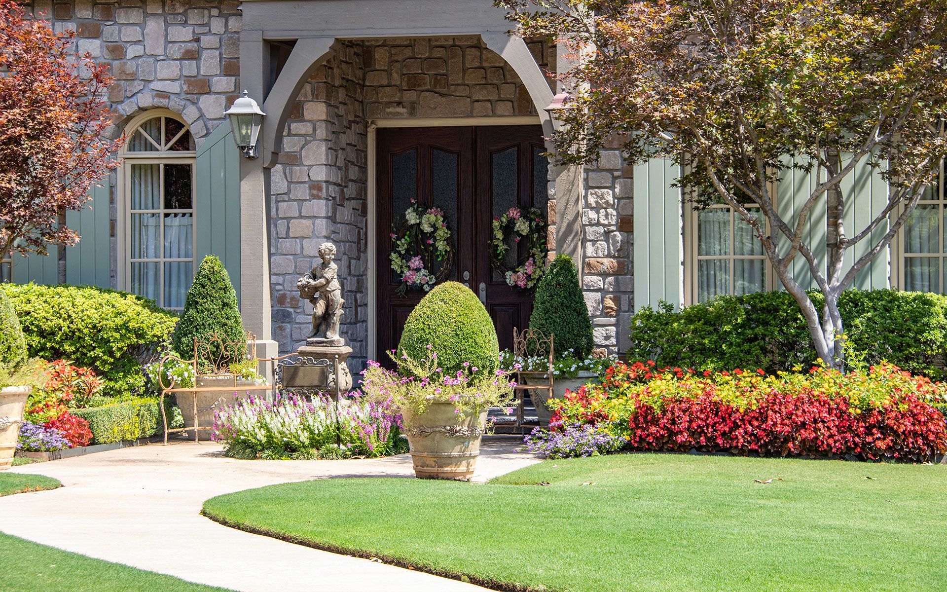 Stone home with a manicured lawn, colorful flowerbeds, and an ornate front door adorned with wreaths.