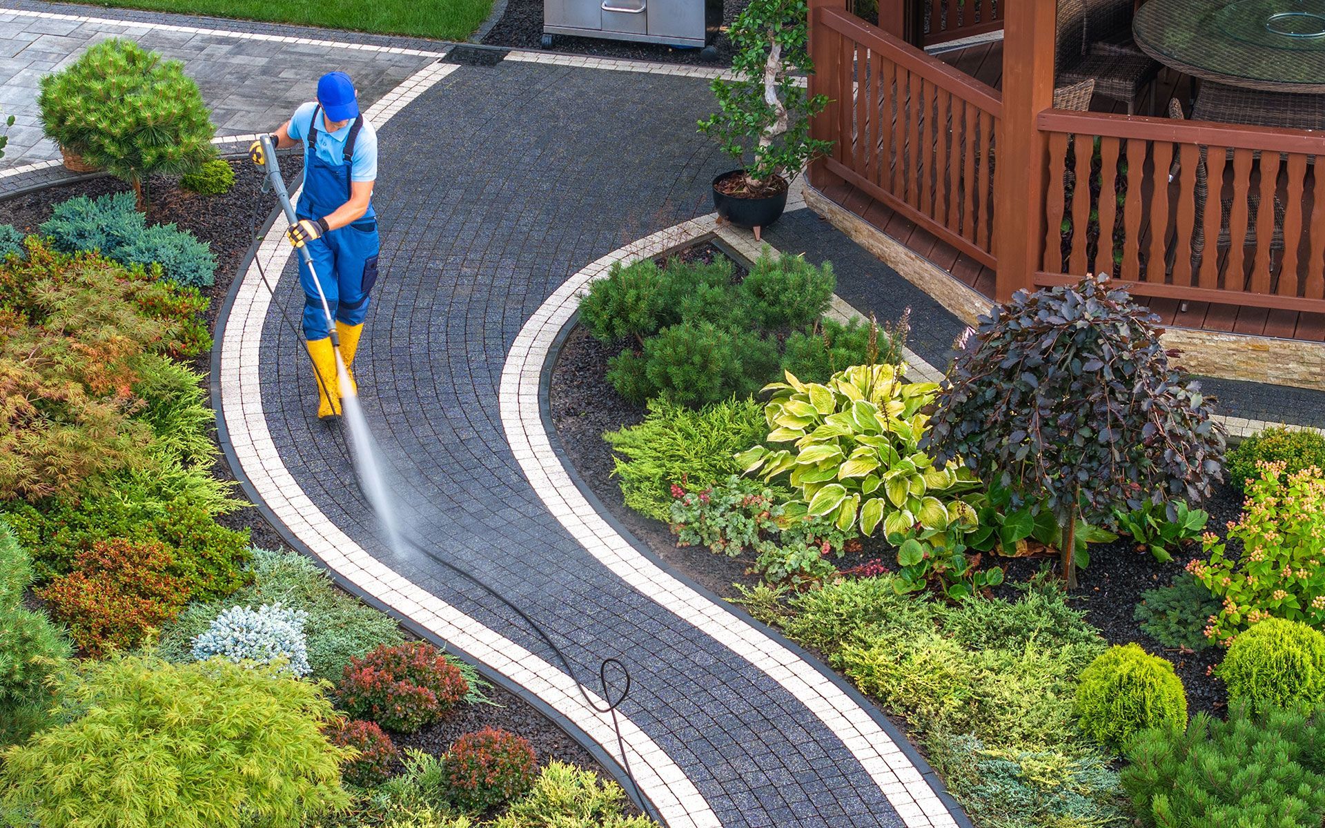 Person power washing a winding black cobblestone path bordered by lush green landscaping.