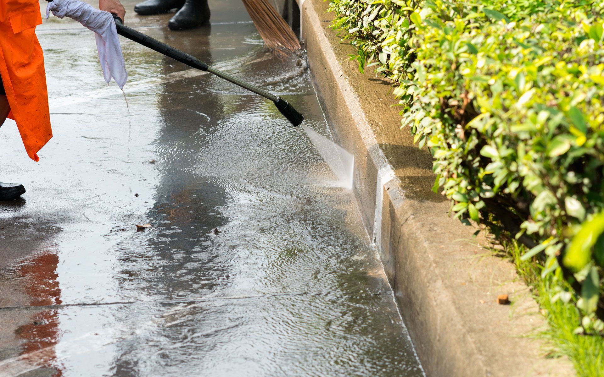 Person in orange robe power washing a sidewalk curb next to green bushes.