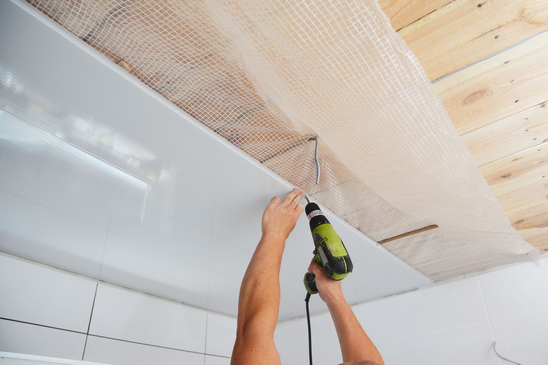 A person uses a power drill to install a ceiling frame with mesh netting in a room under renovation.
