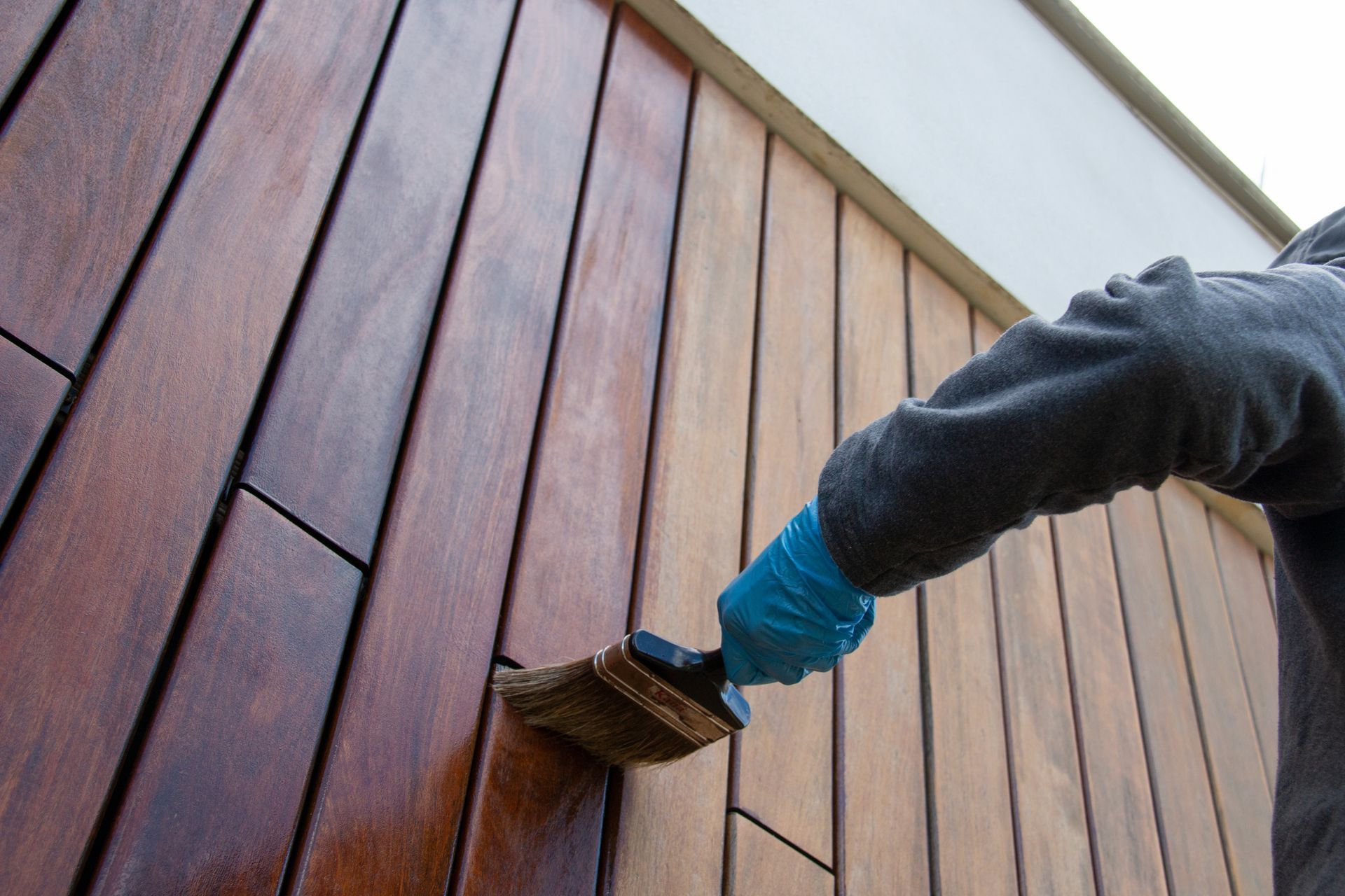 Angled picture of painter with gloves as he brushes oil to wooden cladding on wall.