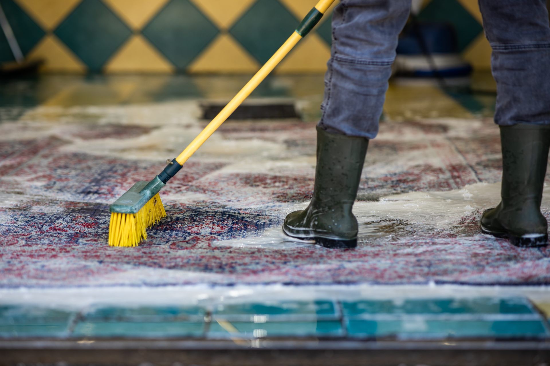 Person scrubbing a wet tiled floor with a yellow brush and squeegee boots visible