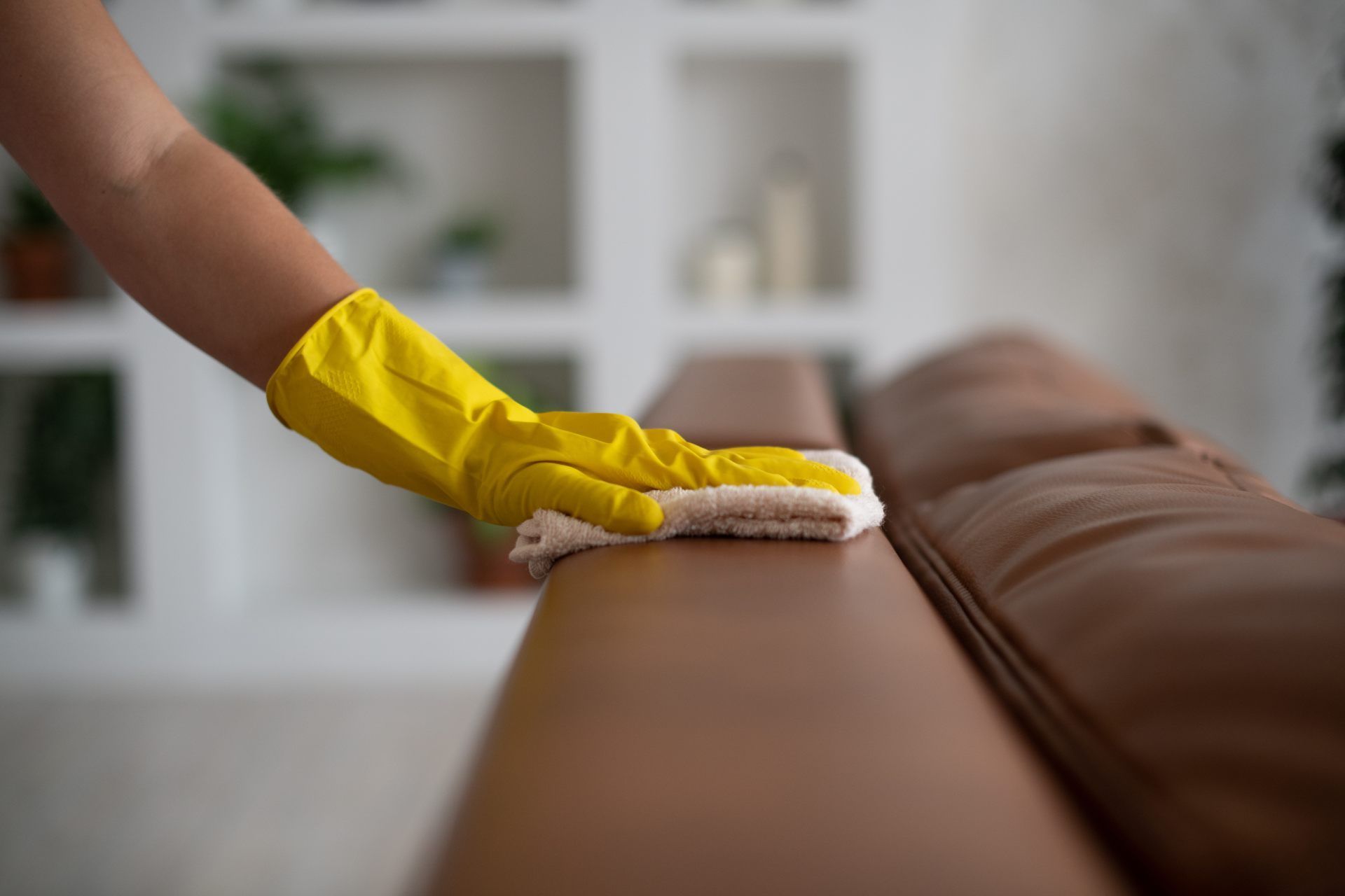 Hand in yellow glove wiping a brown leather couch with a cloth