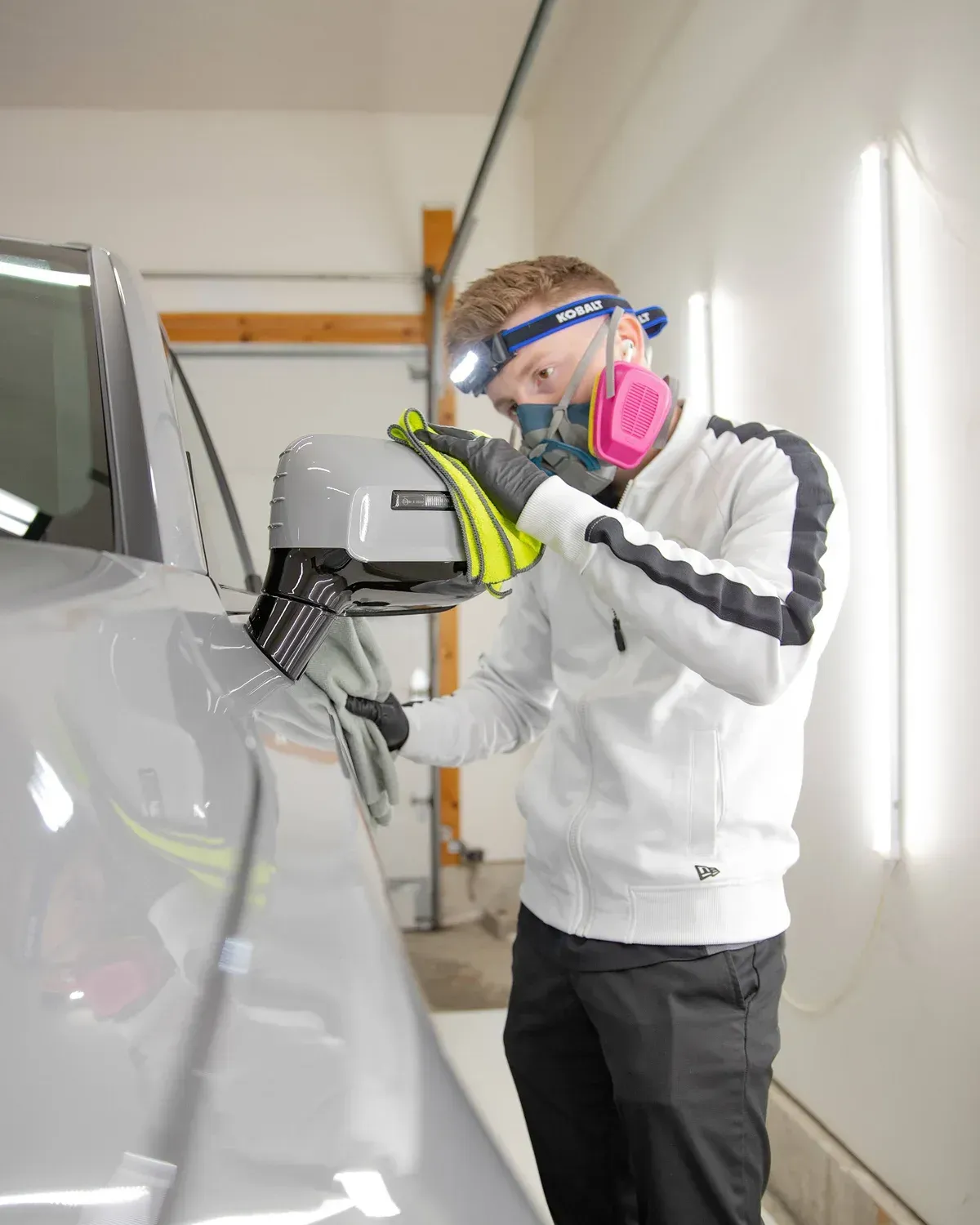 Man cleaning a gray car's side mirror, wearing a respirator, headlamp, and gloves in a garage setting.