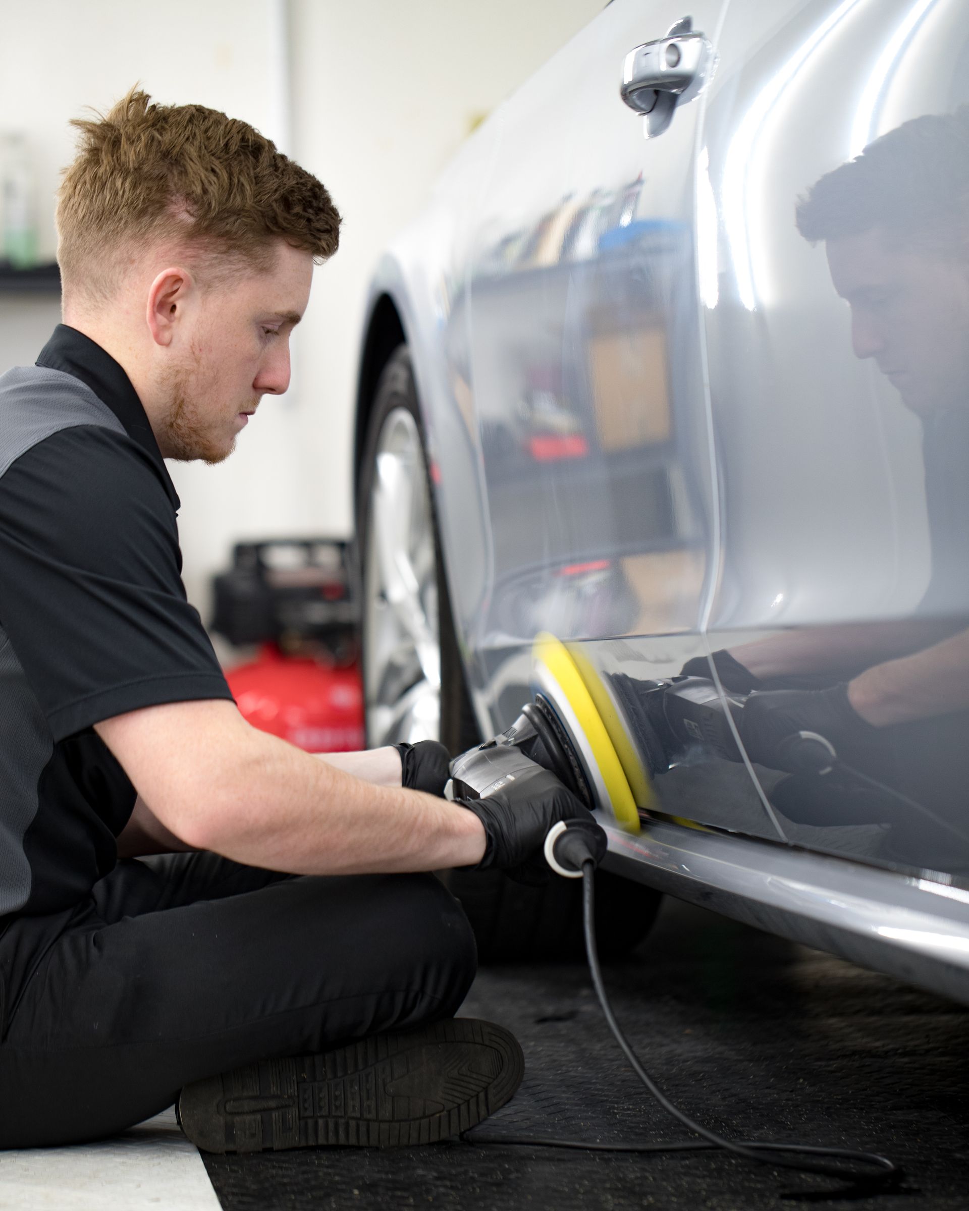 A person polishes a silver car's side panel with a machine. Black shirt, dark gloves, kneeling position.