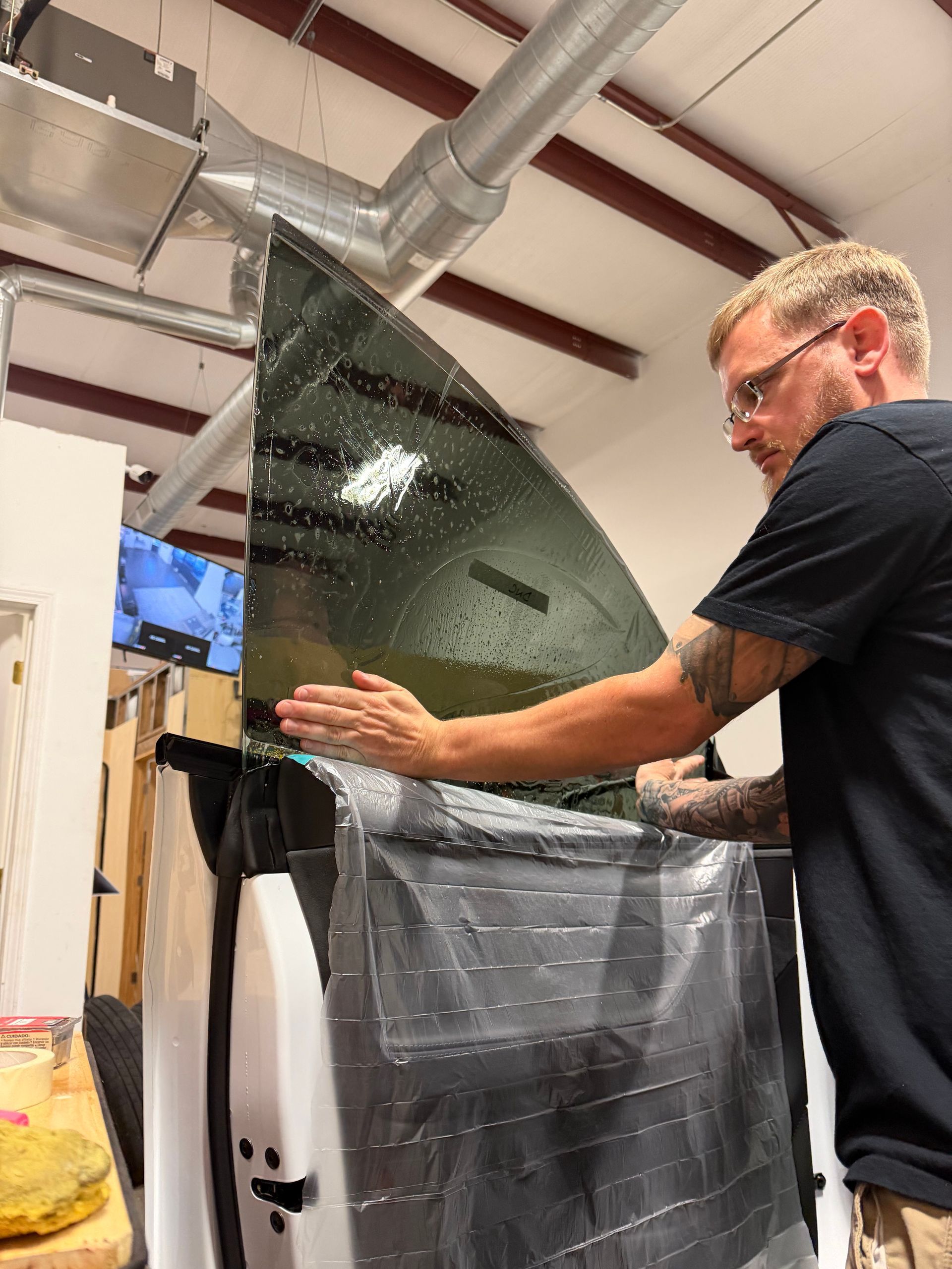 Man applying tint to a car window inside a workshop. He's wearing glasses and has arm tattoos, the window is wet.