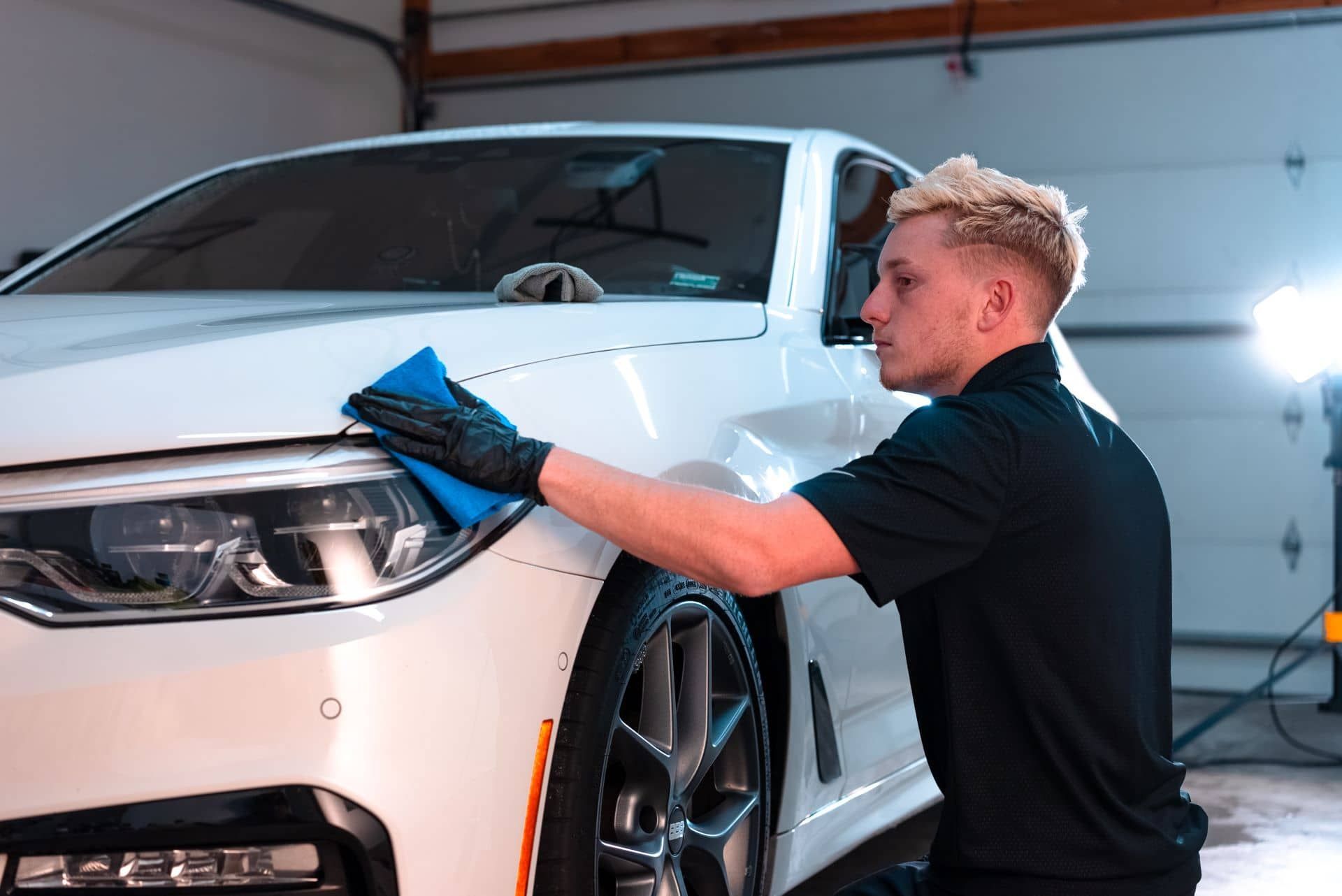 Man in black shirt wiping a white car with a blue cloth in a garage.