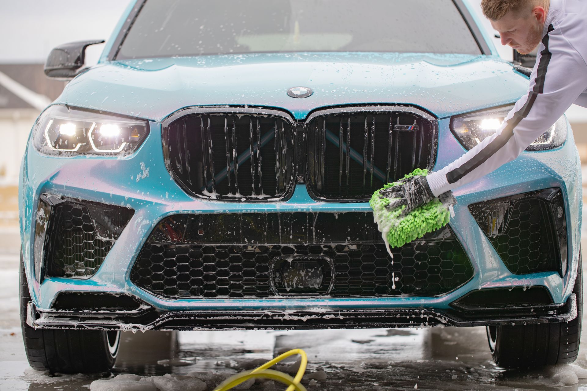 A person washing a teal BMW SUV with a soapy brush outside.