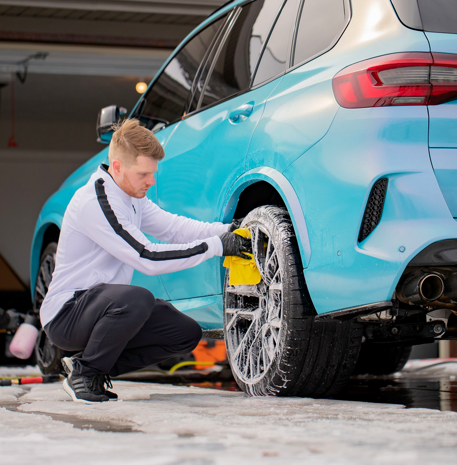 Man washing the tire of a teal SUV with a yellow sponge, outdoors.
