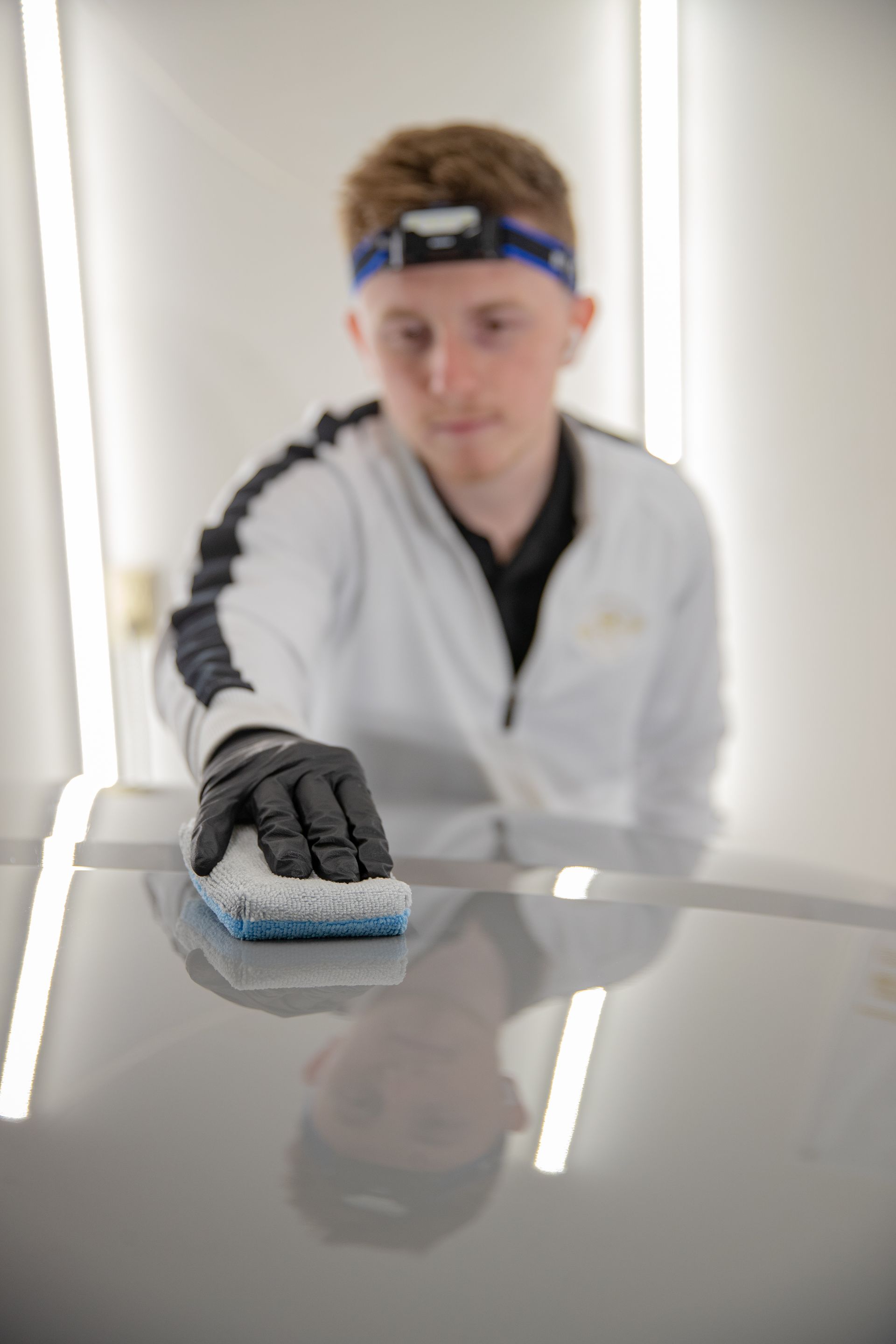 Person in gloves uses a sponge on a car's hood, illuminated by overhead lights.