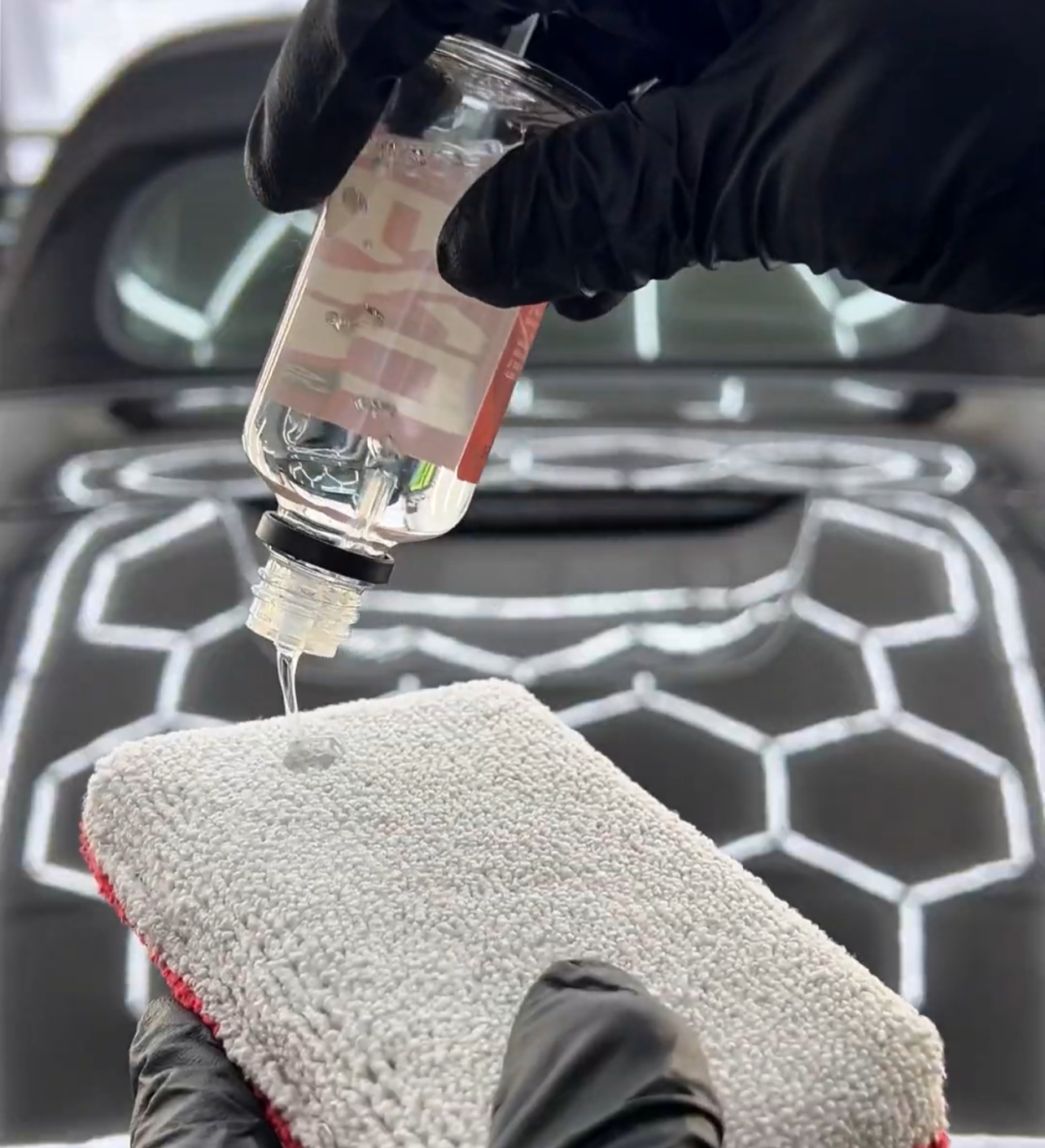 Gloved hand pouring liquid from a bottle onto a microfiber applicator. Car hood in background.