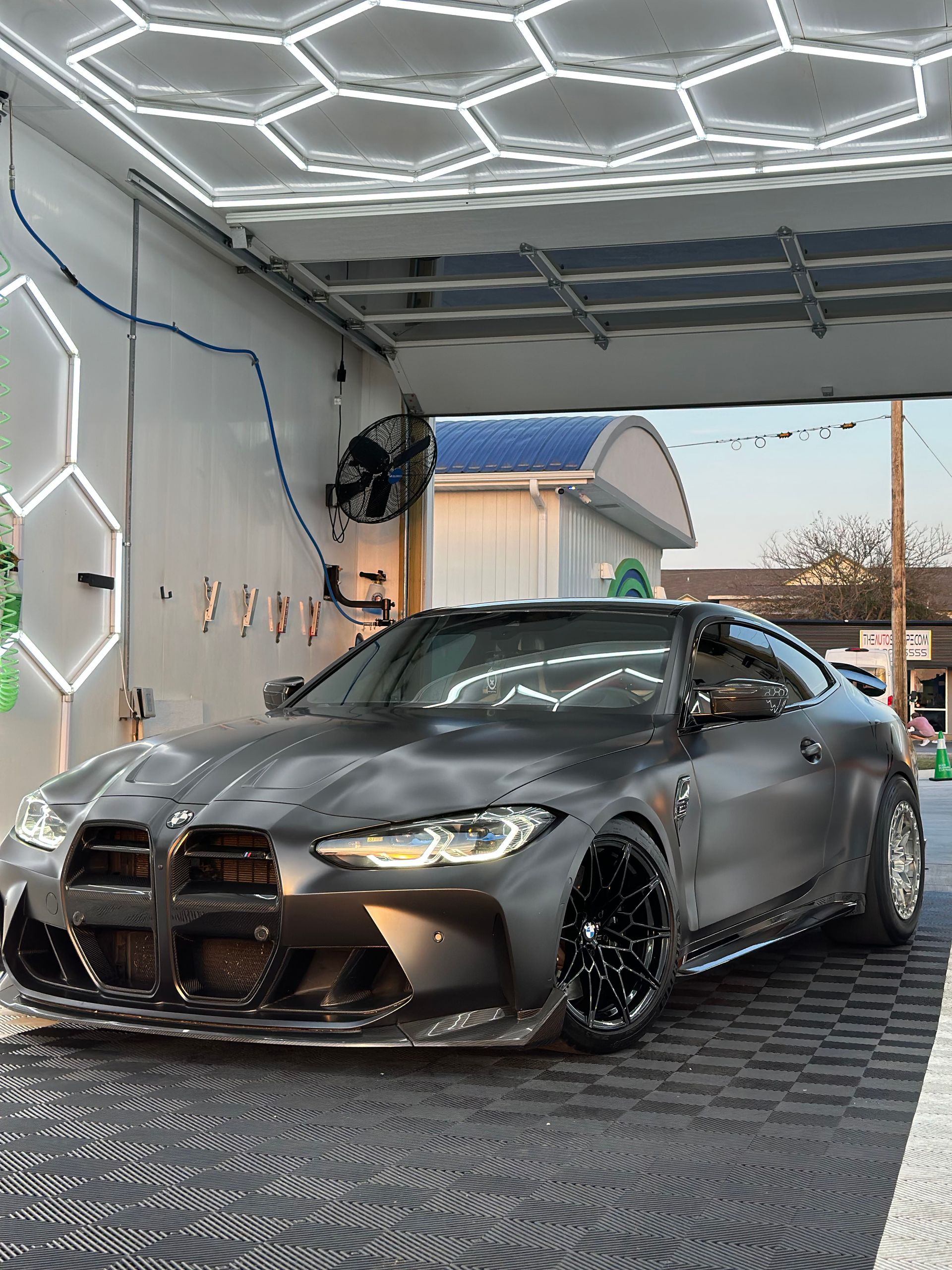Dark gray BMW M4 at a car wash, under hexagonal lights, with black rims.