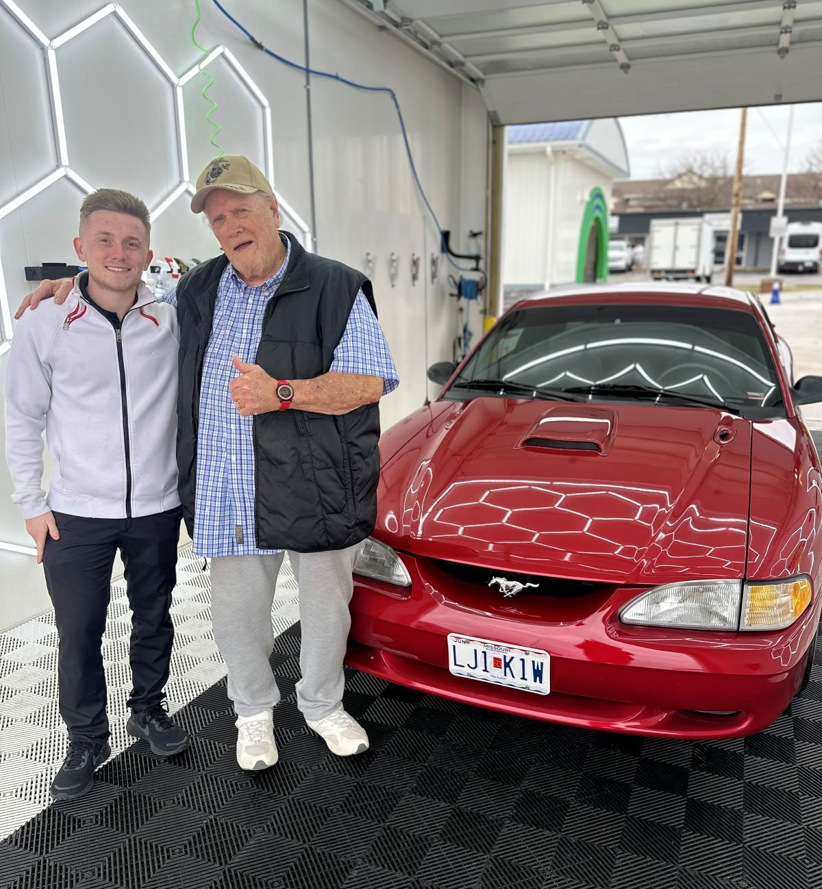 Two men pose next to a red Ford Mustang in a car wash bay. One man gives a thumbs-up.