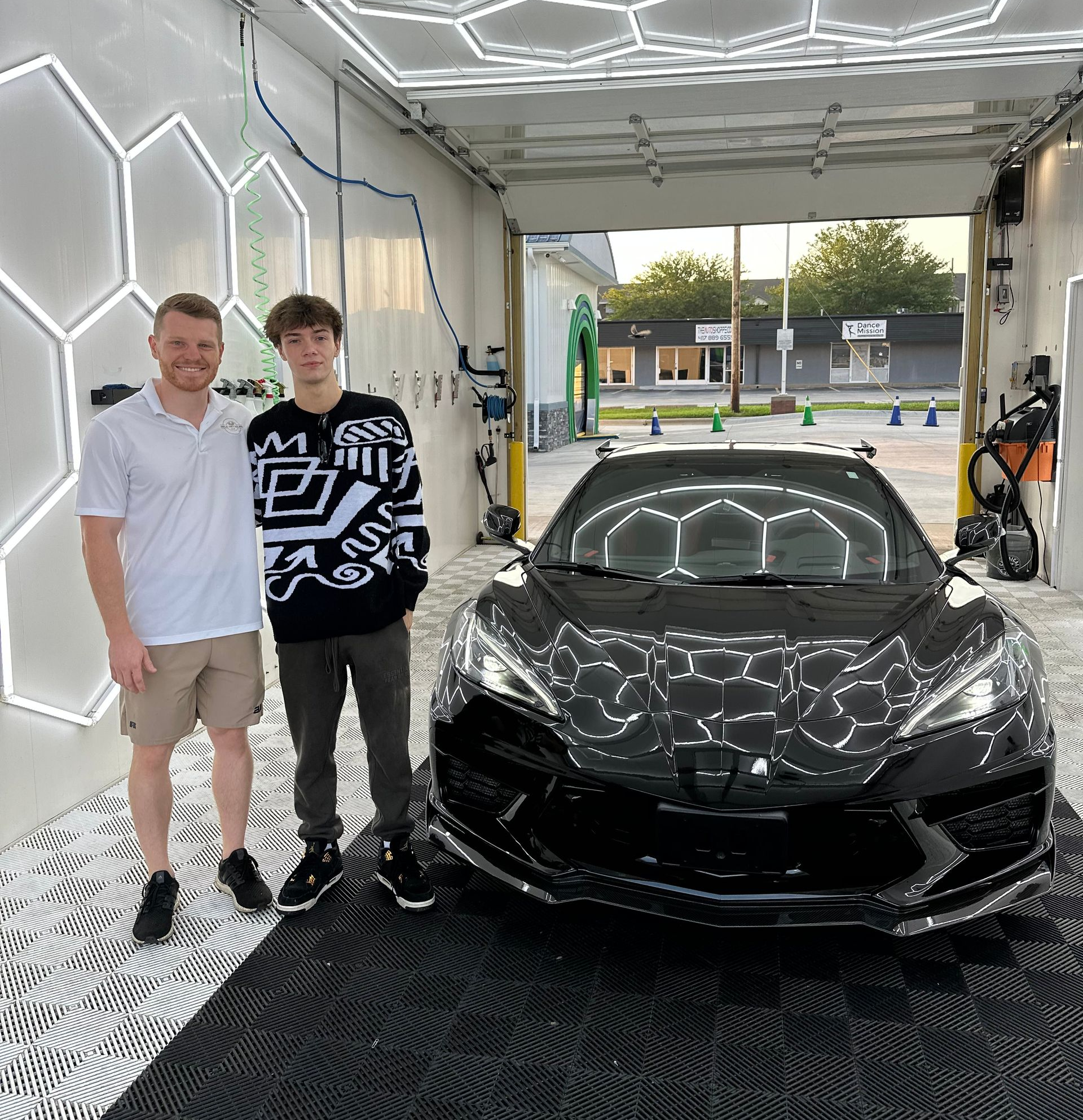 Two people pose with a black sports car in a car wash, under hexagon lights.