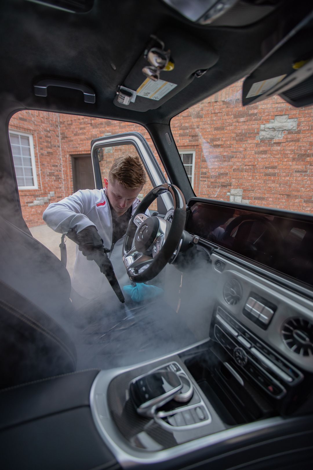 Person steam-cleaning the interior of a luxury car. Steam fills the air; the car sits in front of a brick building.