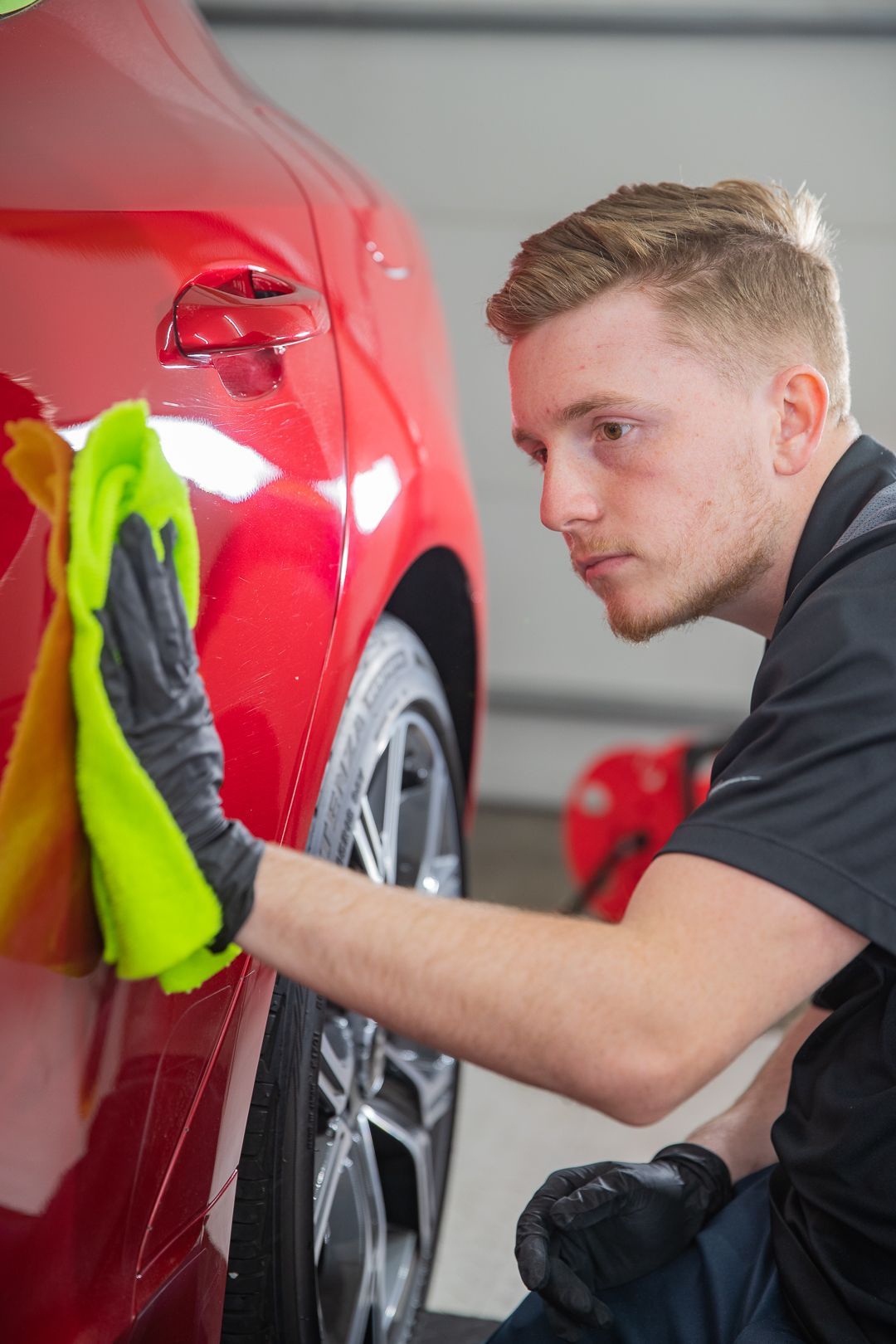 Man in black gloves cleaning a red car with a yellow microfiber cloth.