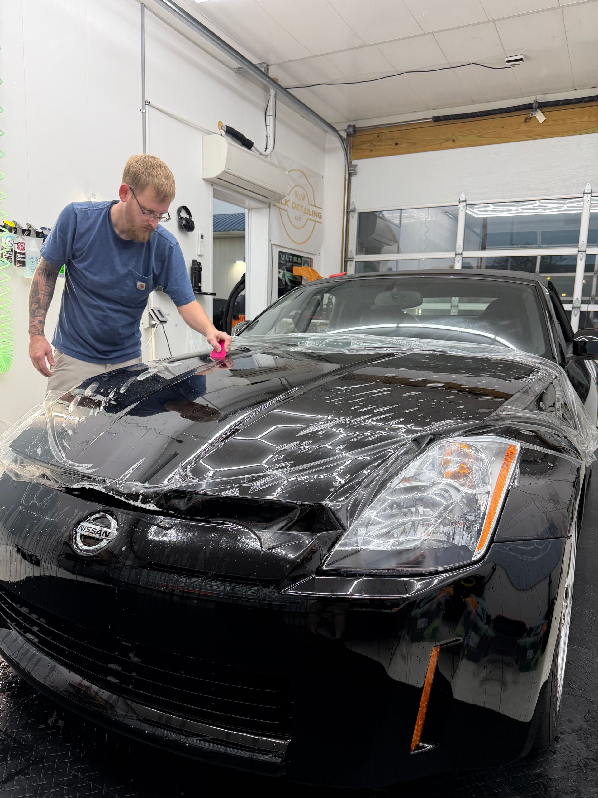 Man washes a black car in a well-lit garage, applying soap with a pink sponge.