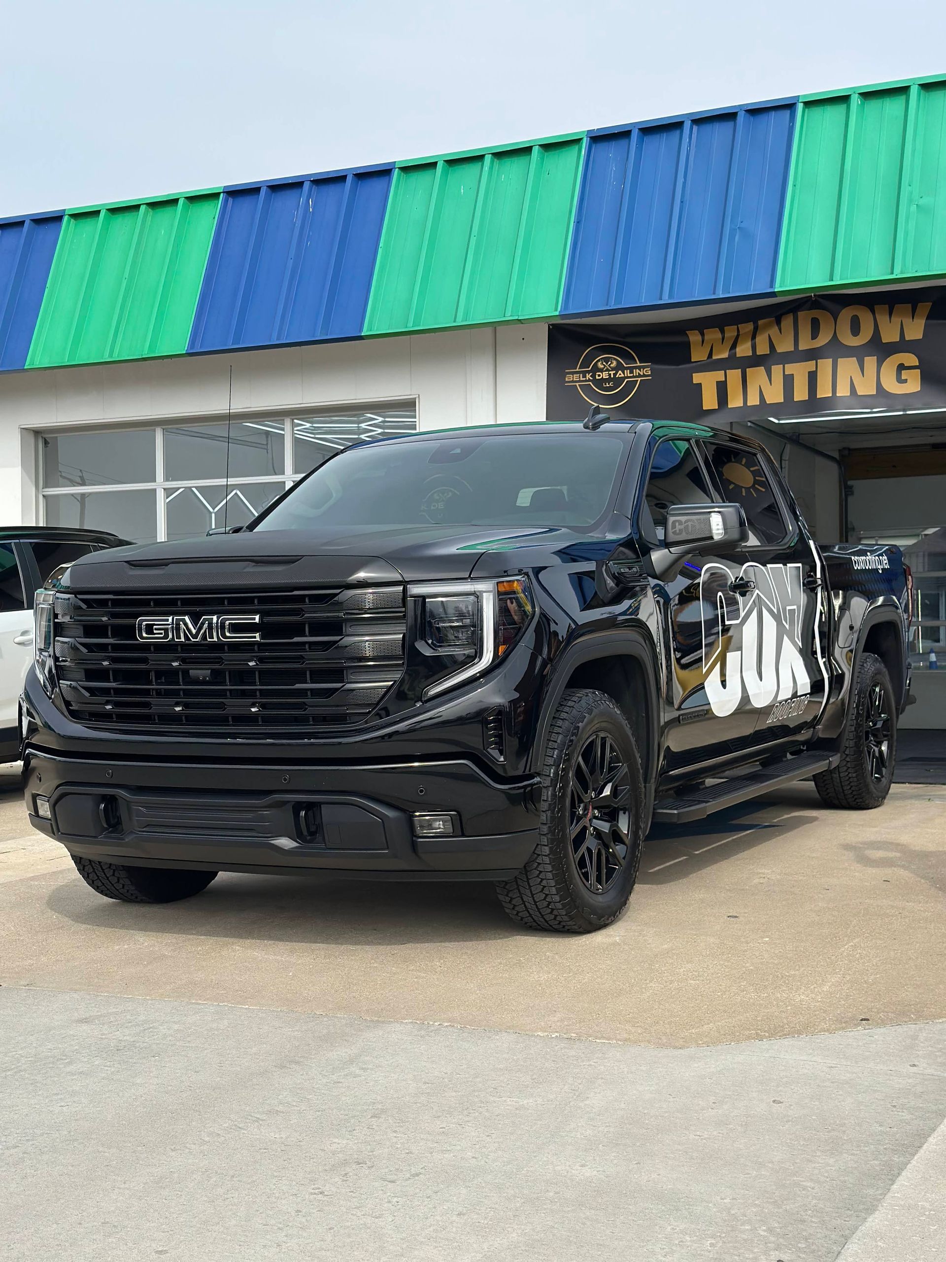 Black GMC truck parked in front of a window tinting shop with a green and blue awning.