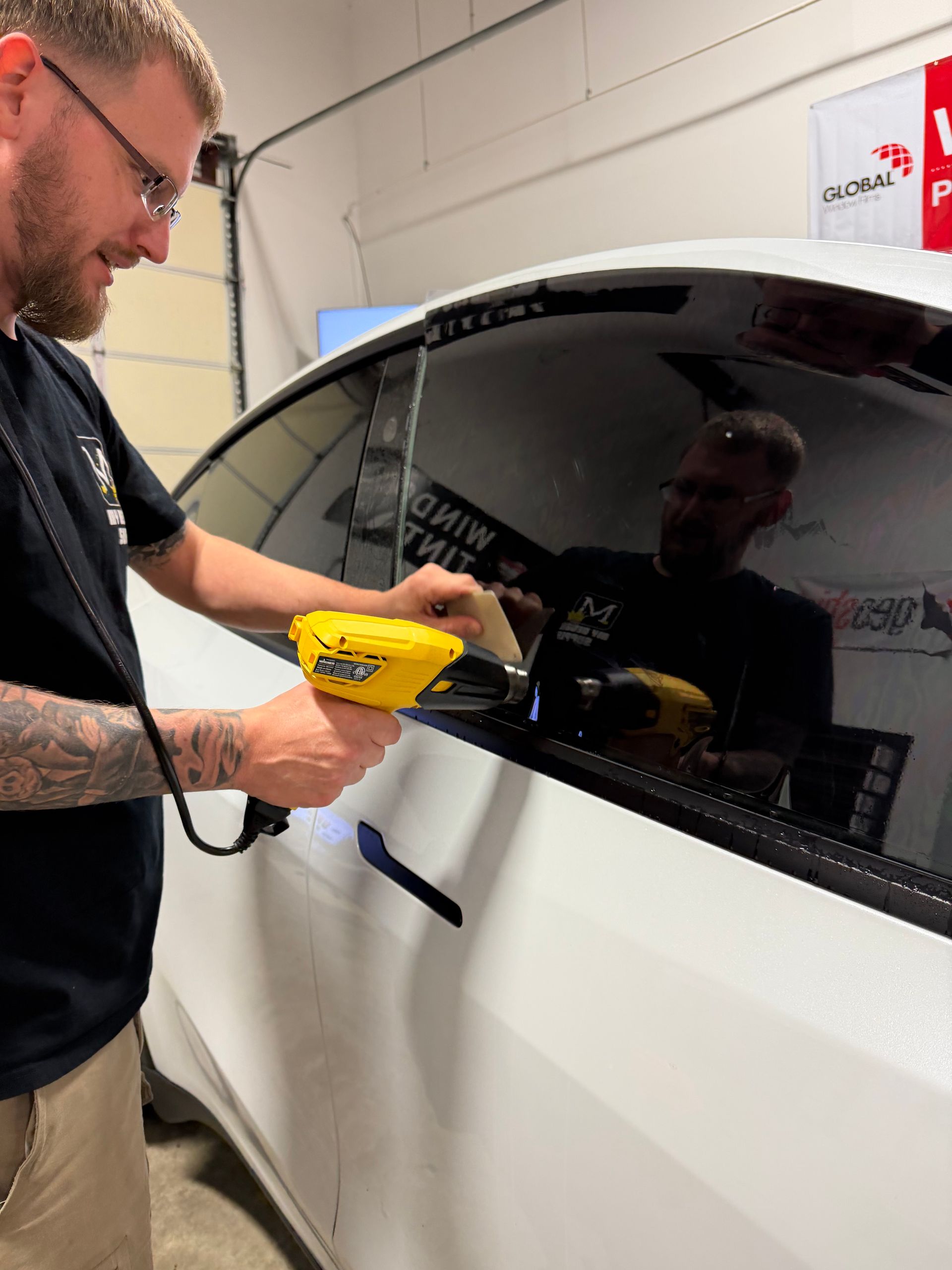 Man uses a heat gun to apply tint to a car window in a garage.
