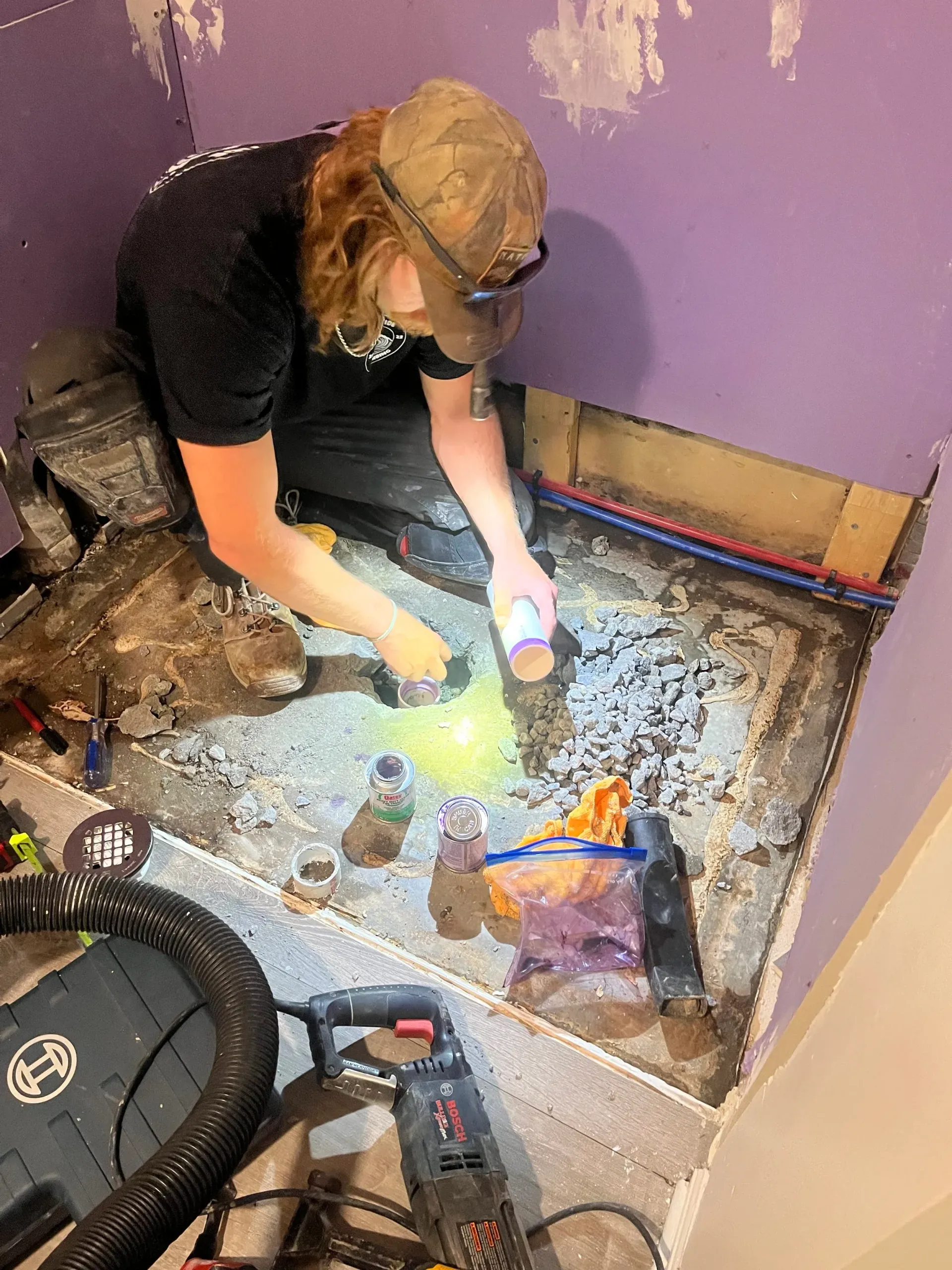Person working on a bathroom floor, using a flashlight. Purple walls, exposed pipes and debris visible.